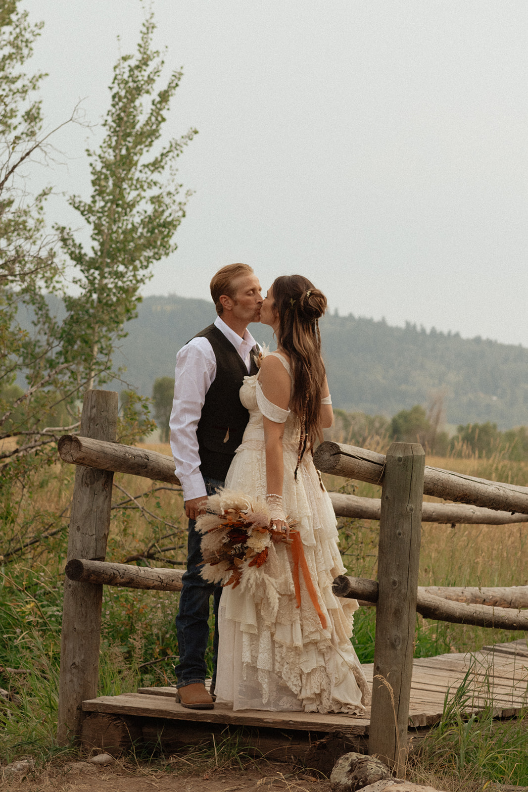 A couple dressed in wedding attire stands on a wooden bridge. The man, in a vest, embraces and kisses the woman, dressed in a flowing white gown. They are surrounded by a natural outdoor setting for their wedding at preston branch