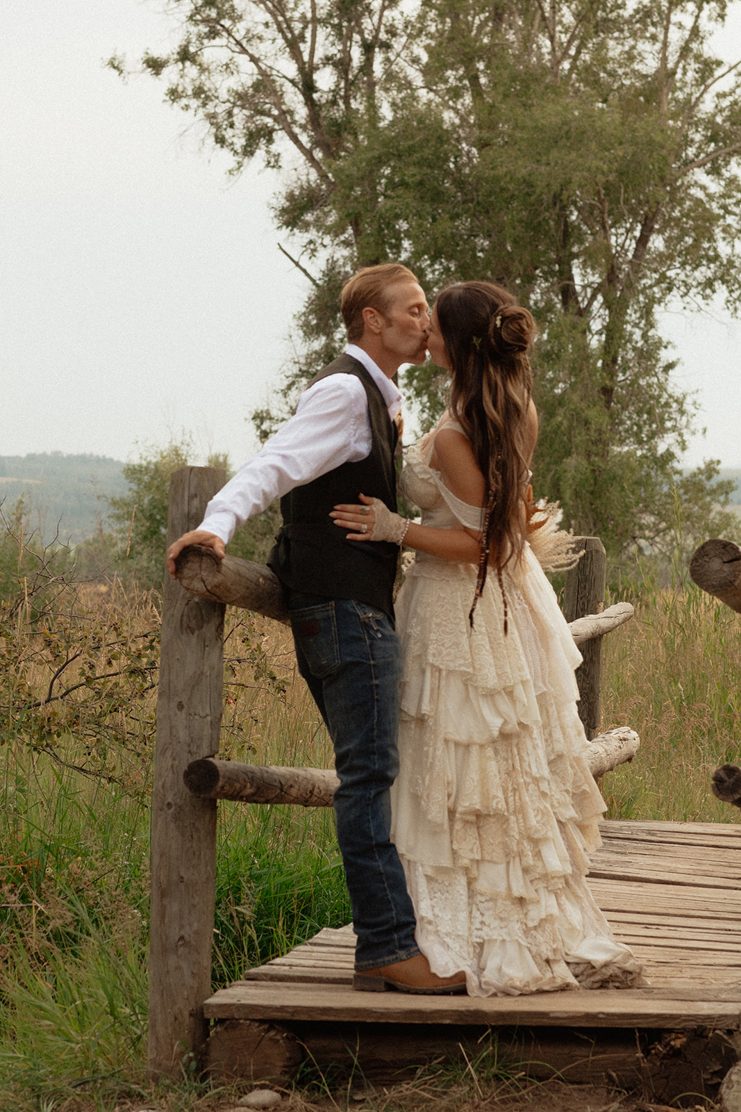 A couple dressed in wedding attire stands on a wooden bridge. The man, in a vest, embraces and kisses the woman, dressed in a flowing white gown. They are surrounded by a natural outdoor setting for their wedding at preston branch