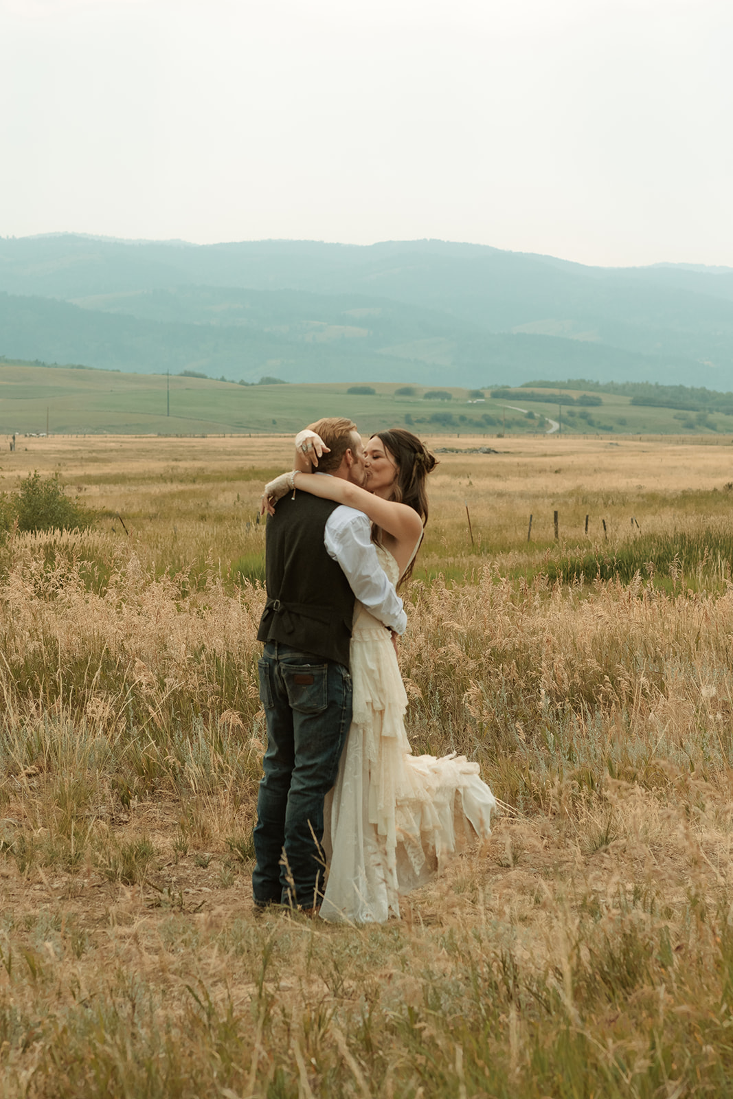 A couple dressed in wedding attire stands on a wooden bridge. The man, in a vest, embraces and kisses the woman, dressed in a flowing white gown. They are surrounded by a natural outdoor setting for their wedding at preston branch