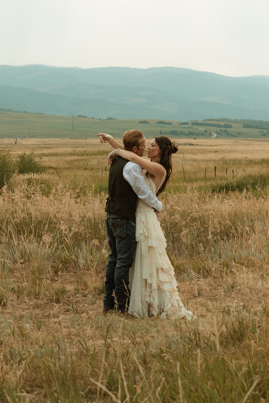 A couple dressed in wedding attire stands on a wooden bridge. The man, in a vest, embraces and kisses the woman, dressed in a flowing white gown. They are surrounded by a natural outdoor setting for their wedding at preston branch