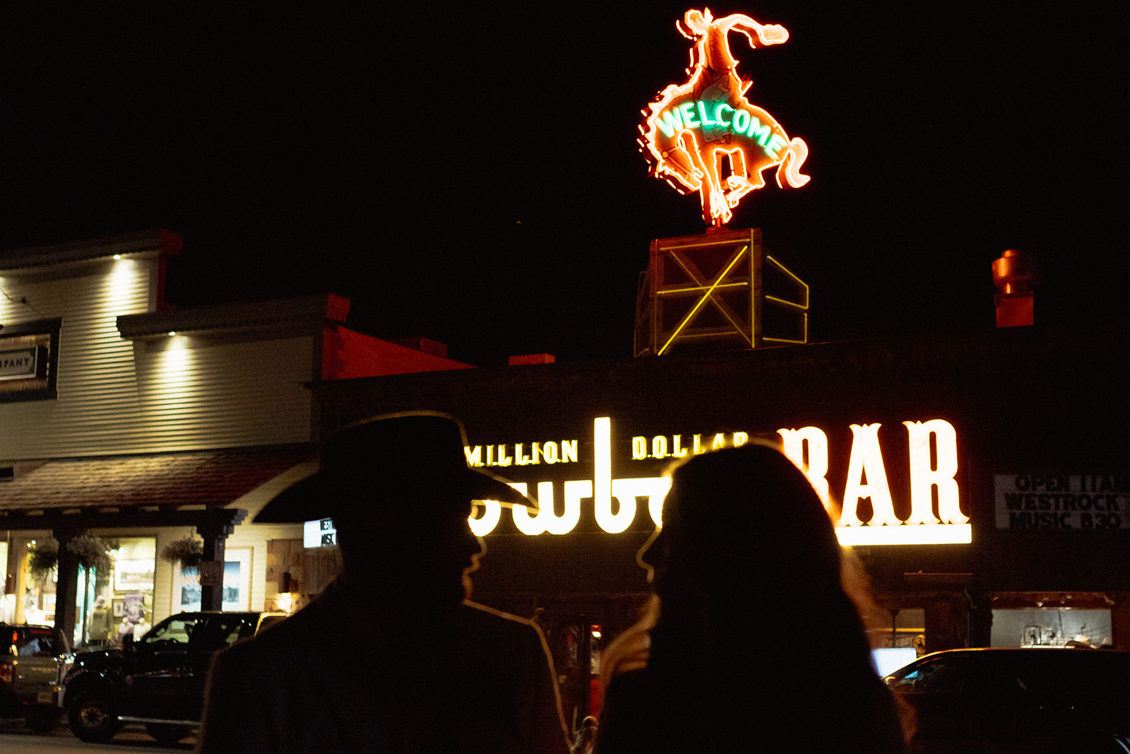 Person standing on a road at night, illuminated by a neon sign for the Cowboy Bar. Nearby building lights are also visible in the background.