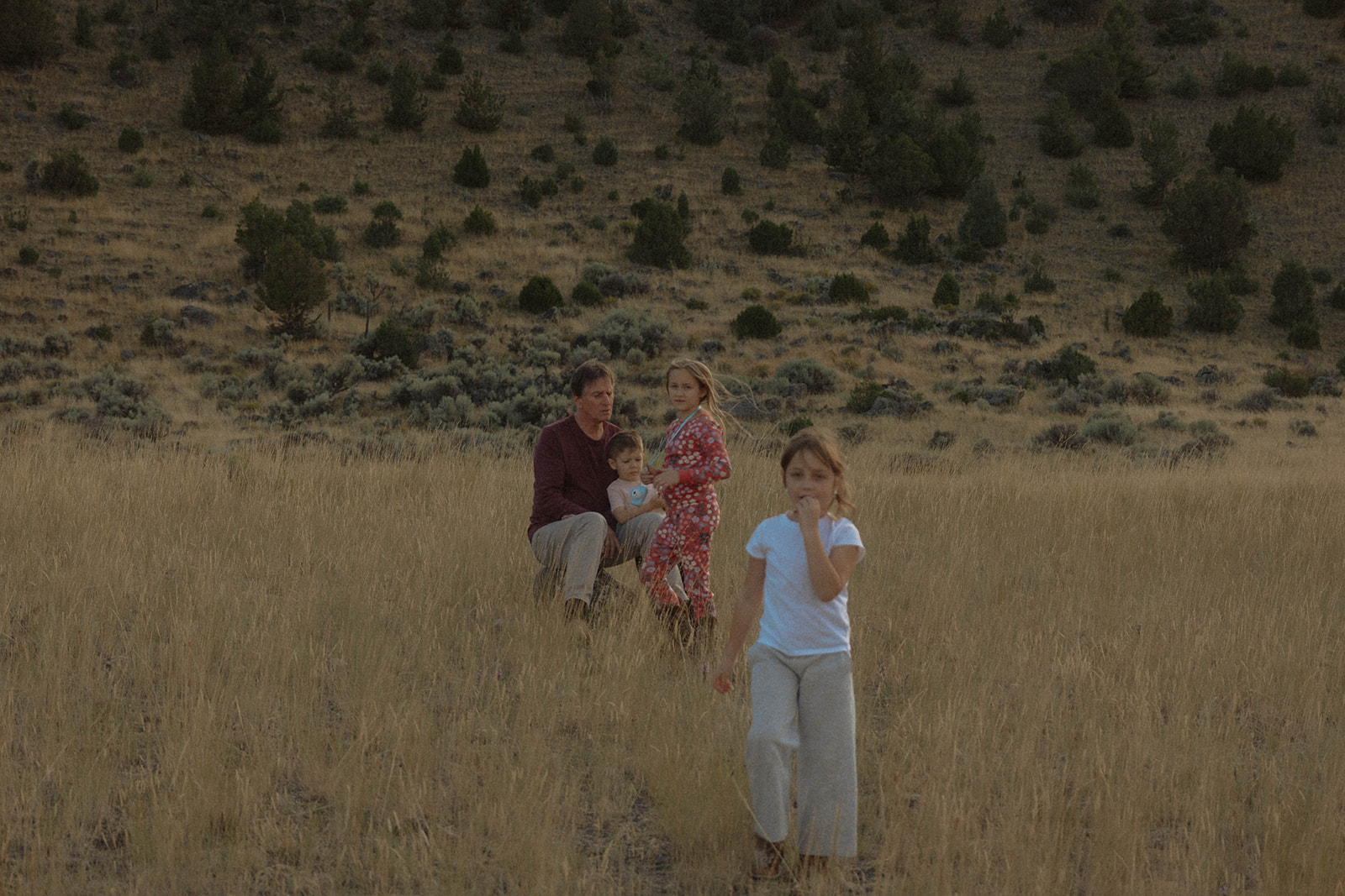 A young boy stands in the foreground in a grassy field, while a couple dressed in formal attire is blurry in the background with mountainous landscape. | children at your wedding