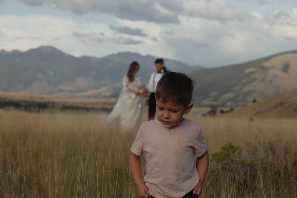 A young boy stands in the foreground in a grassy field, while a couple dressed in formal attire is blurry in the background with mountainous landscape. | children at your wedding