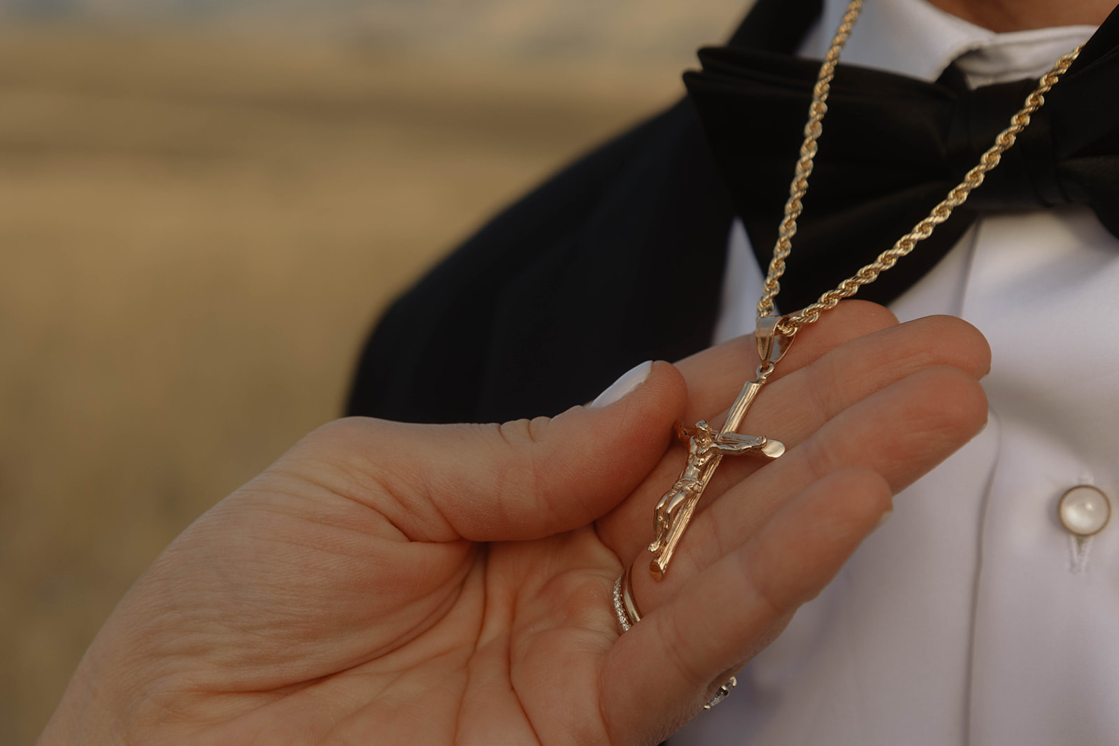 A hand holds a gold crucifix on a chain around a person’s neck, who is dressed in a black bow tie and white shirt.