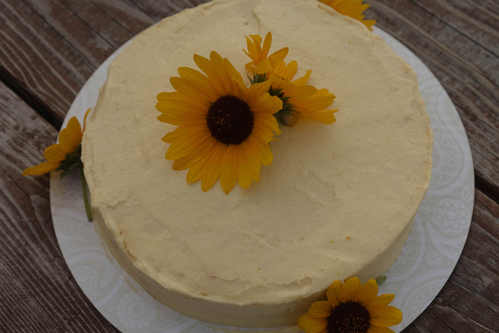 A round cake with smooth frosting, topped with three yellow sunflowers, is placed on a white plate.