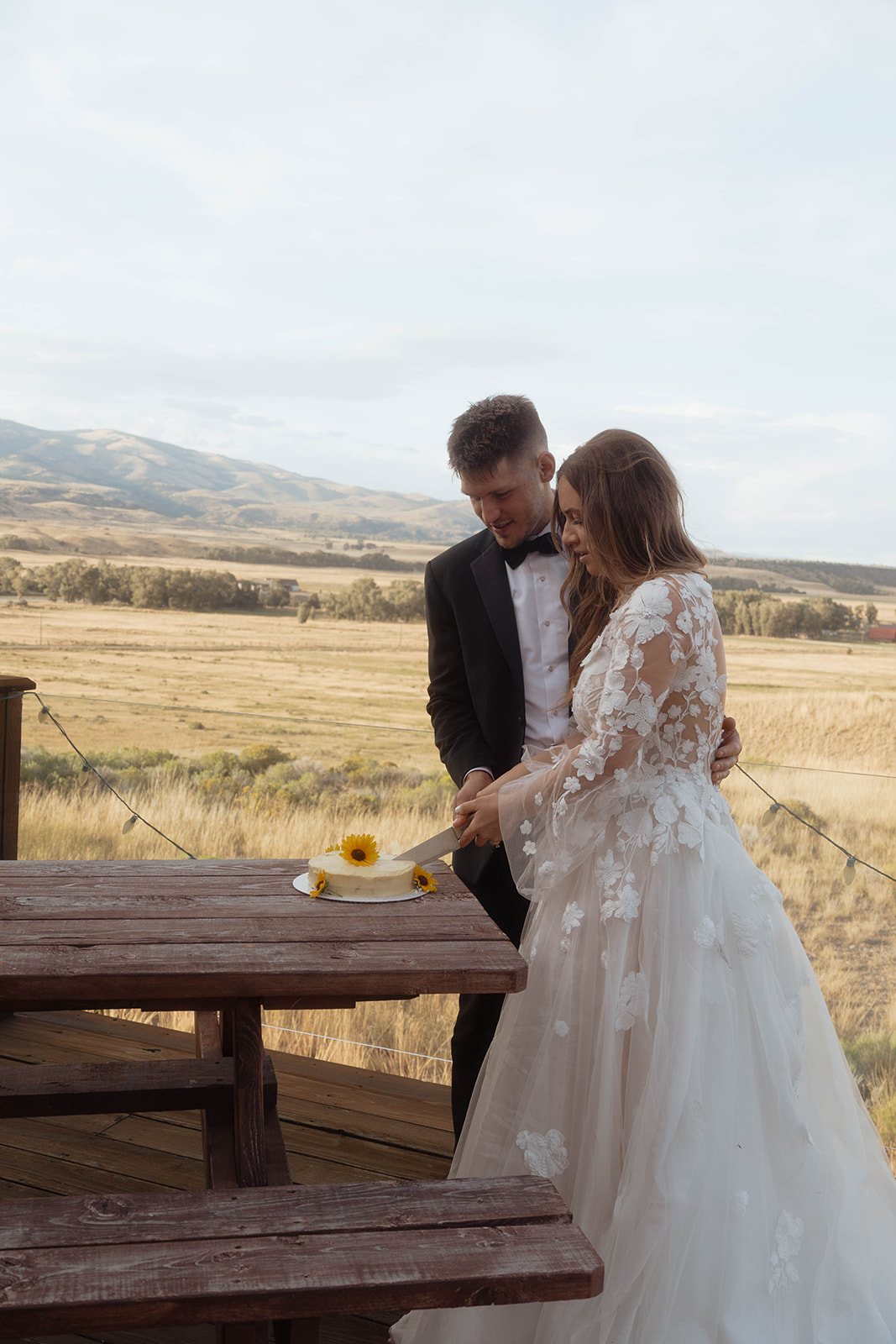 A bride and groom, wearing a wedding dress and tuxedo respectively, stand together outdoors cutting a cake on a wooden table with a field and mountains in the background.