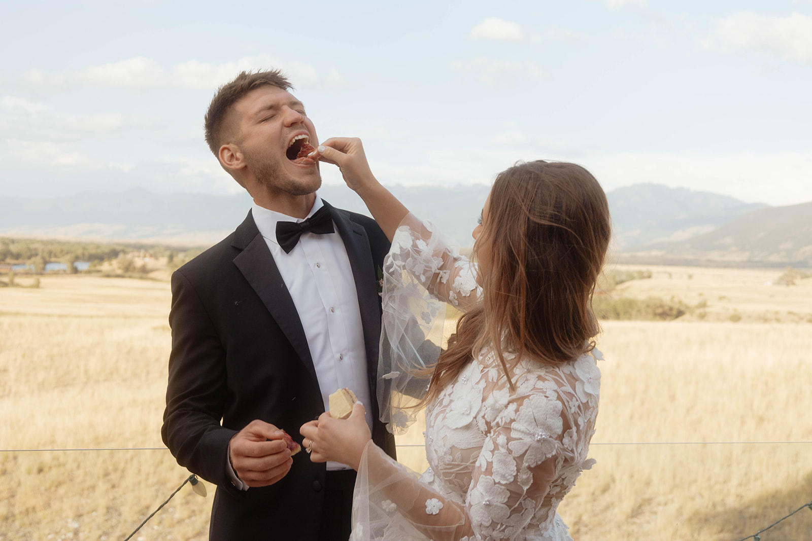 A bride and groom, wearing a wedding dress and tuxedo respectively, stand together outdoors cutting a cake on a wooden table with a field and mountains in the background.