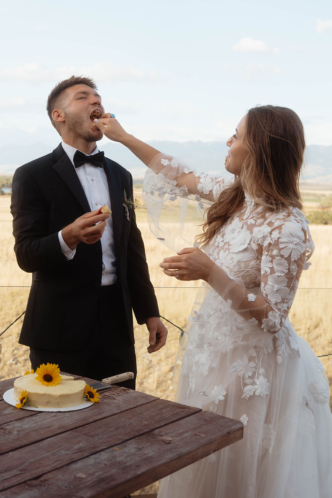 A bride and groom, wearing a wedding dress and tuxedo respectively, stand together outdoors cutting a cake on a wooden table with a field and mountains in the background.