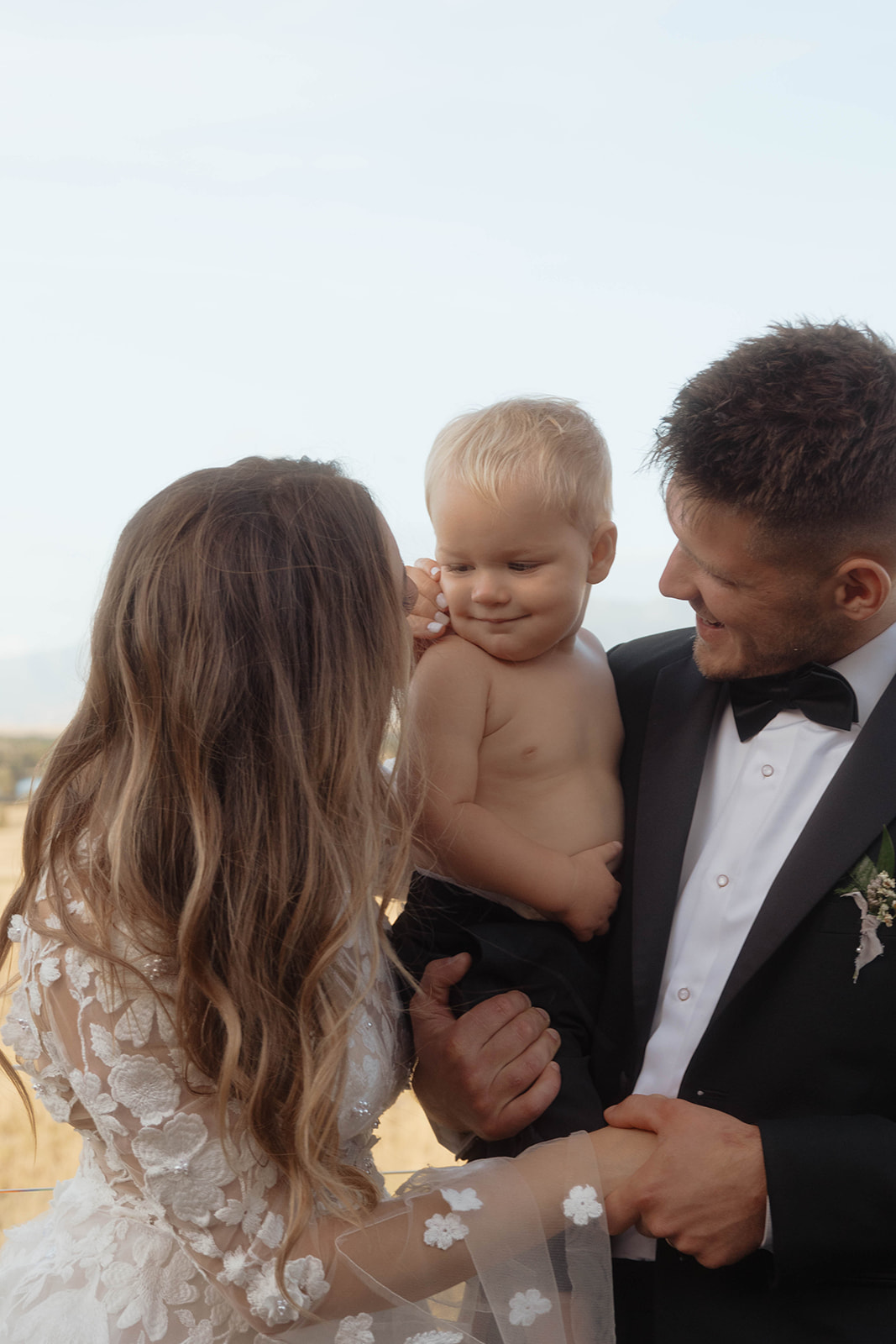 A couple in formal attire stands outdoors, smiling at a shirtless baby the man is holding. | Children at your wedding