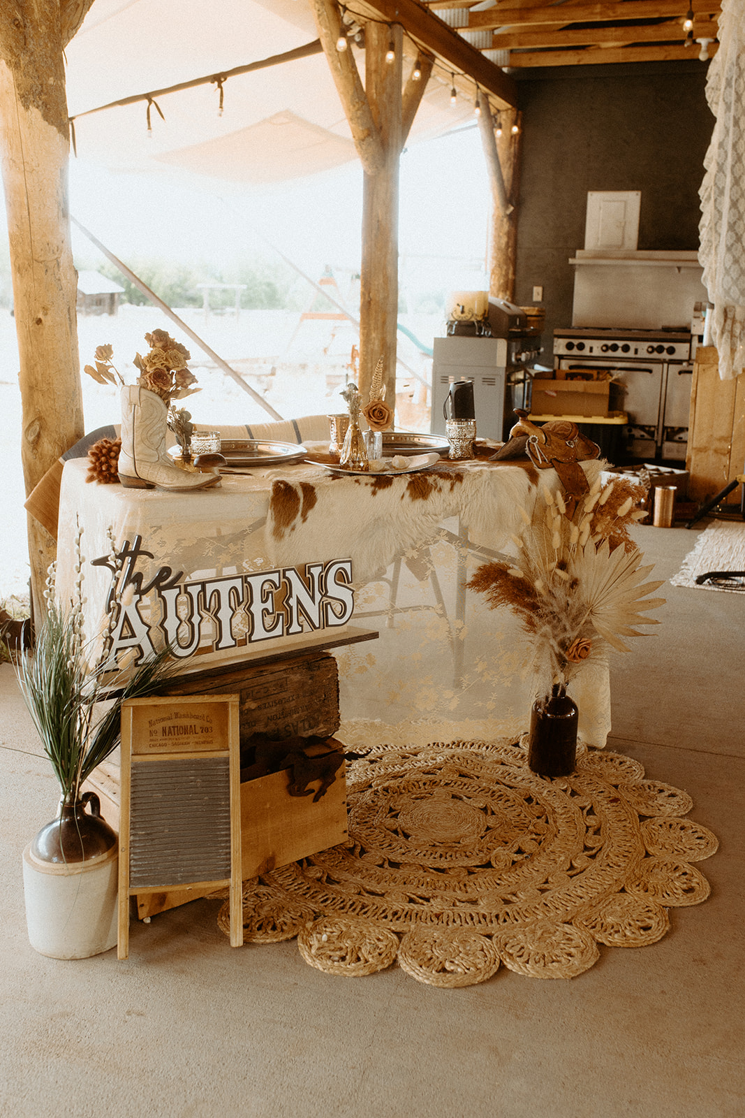 A rustic setup featuring a decorated wooden counter with a sign reading "Austens." The counter is adorned with dried plants, and nearby, there are potted plants and a rug on the floor. A kitchen area is visible in the background.