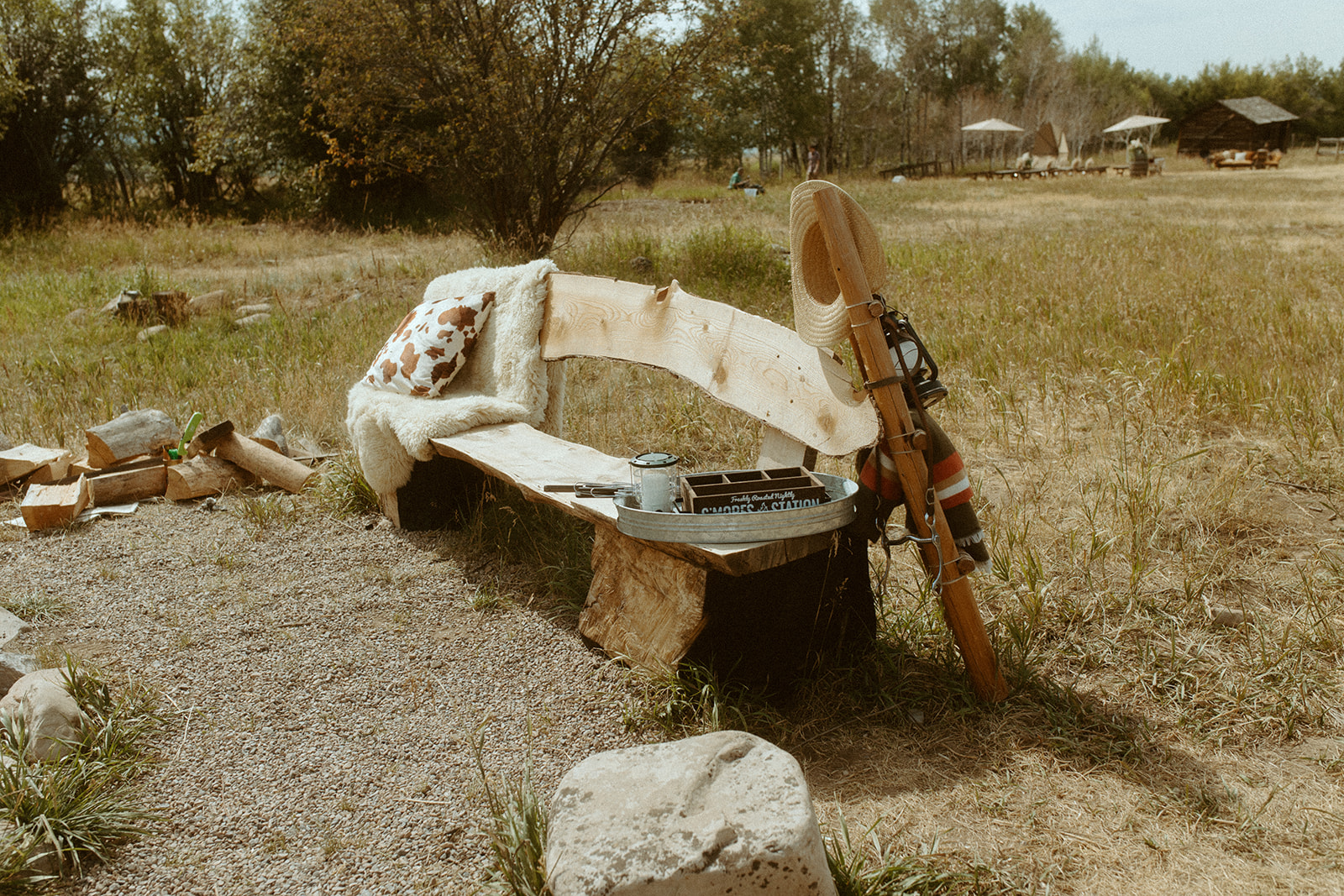 A rustic wooden bench with a blanket and pillow on it sits in a grassy field. Nearby, a guitar is leaning against the bench and some books and a cup of tea are placed on the ground.