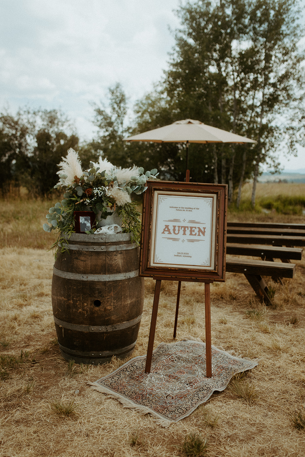 An outdoor wedding setup includes a Welcome sign on an easel next to a decorated wine barrel, flanked by trees and wooden benches.