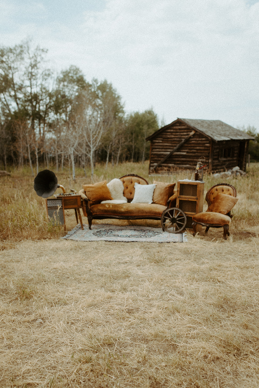 A rustic outdoor scene with a vintage brown sofa, two matching chairs, a gramophone, and a wooden shelf set on a rug in a grassy field with a wooden cabin in the background.