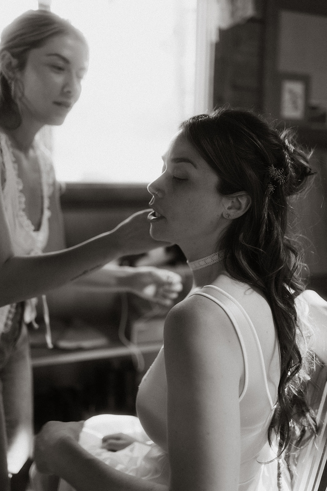 A woman, in a white dress, sits and has her makeup done by another woman in a casual outfit. The setting appears to be indoors and lit by natural light for her wedding at preston ranch