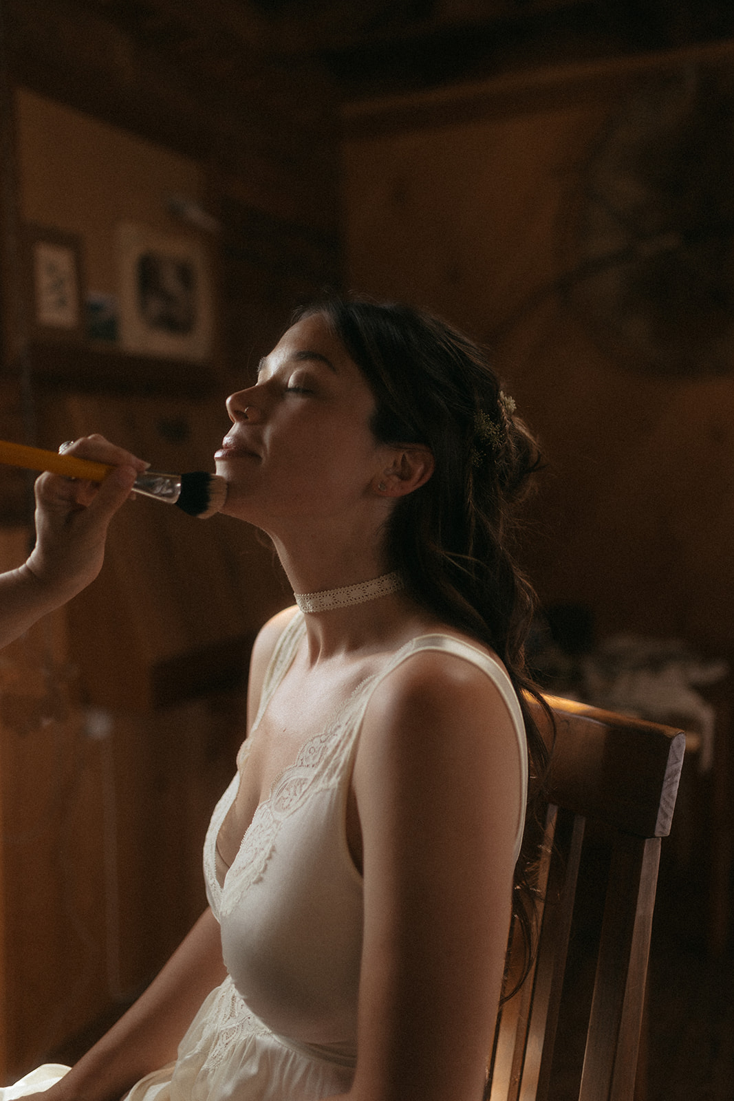 A woman, in a white dress, sits and has her makeup done by another woman in a casual outfit. The setting appears to be indoors and lit by natural light for her wedding at preston ranch