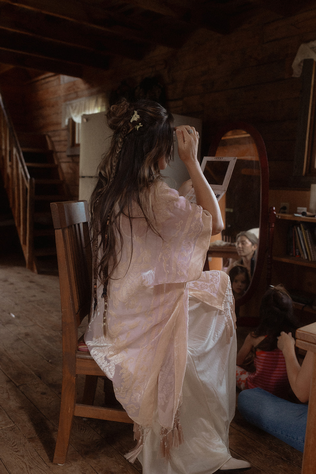 A woman, in a white dress, sits and has her makeup done by another woman in a casual outfit. The setting appears to be indoors and lit by natural light for her wedding at preston ranch