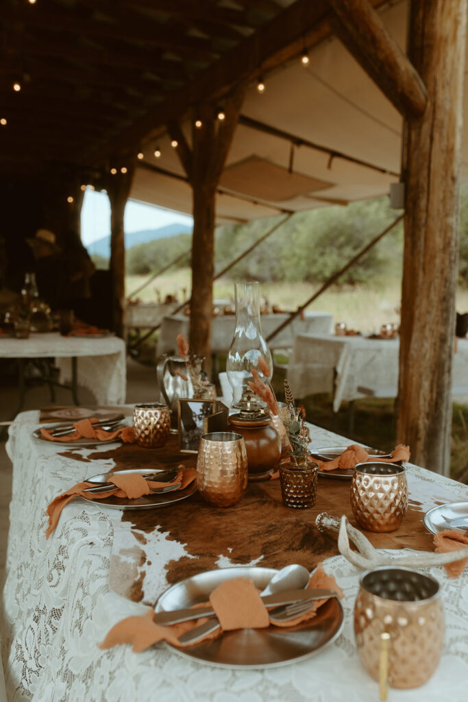 A rustic outdoor dining table setup with lace tablecloth, copper mugs, and earth-toned napkins under a wooden canopy with string lights, surrounded by other tables and scenic natural background.