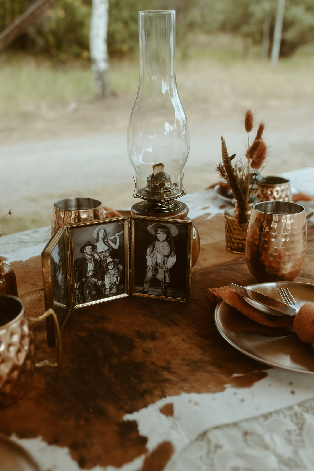 A rustic outdoor dining table setup with lace tablecloth, copper mugs, and earth-toned napkins under a wooden canopy with string lights, surrounded by other tables and scenic natural background.