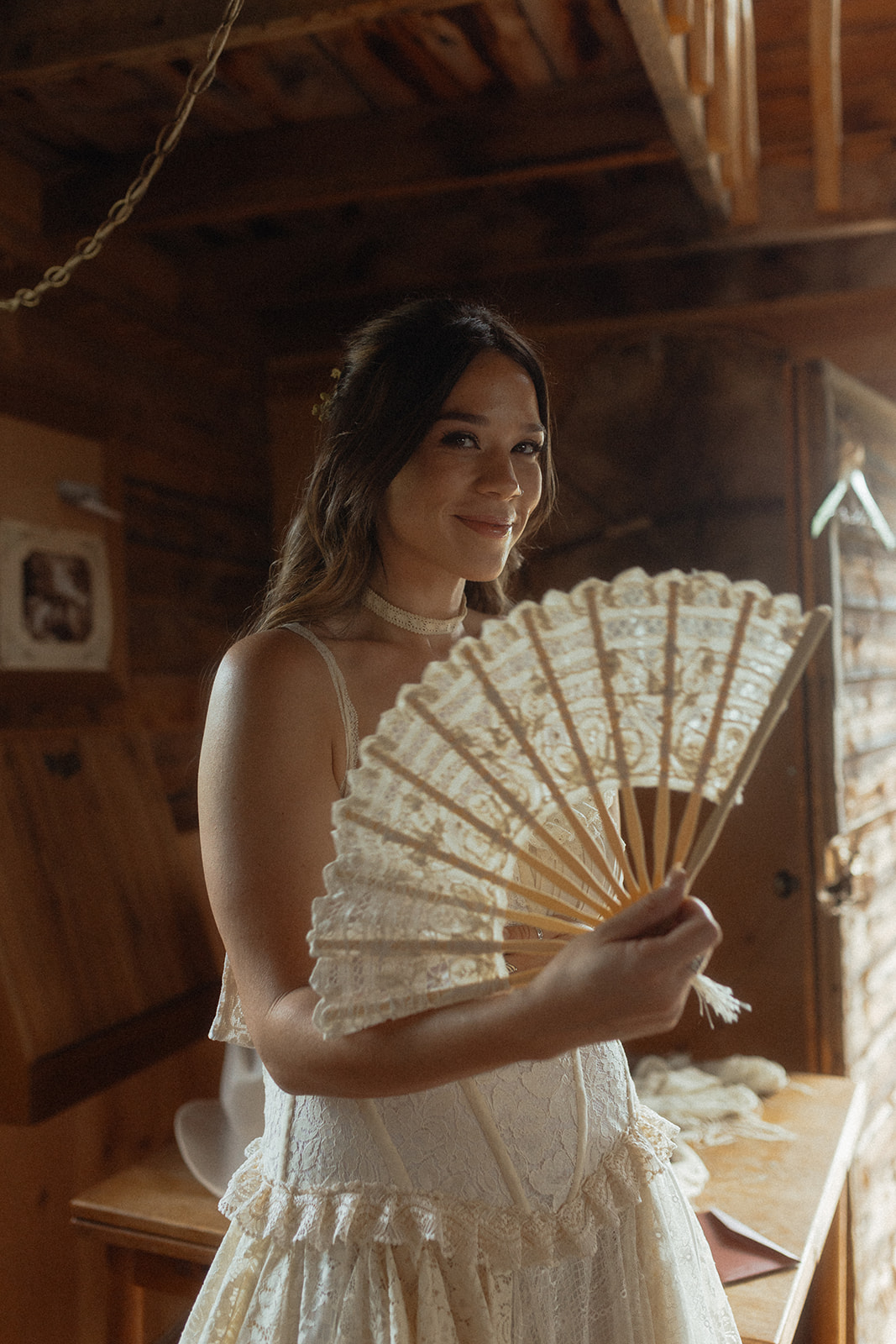 A woman in a white lace dress holds an intricate lace fan while standing in a rustic wooden room.