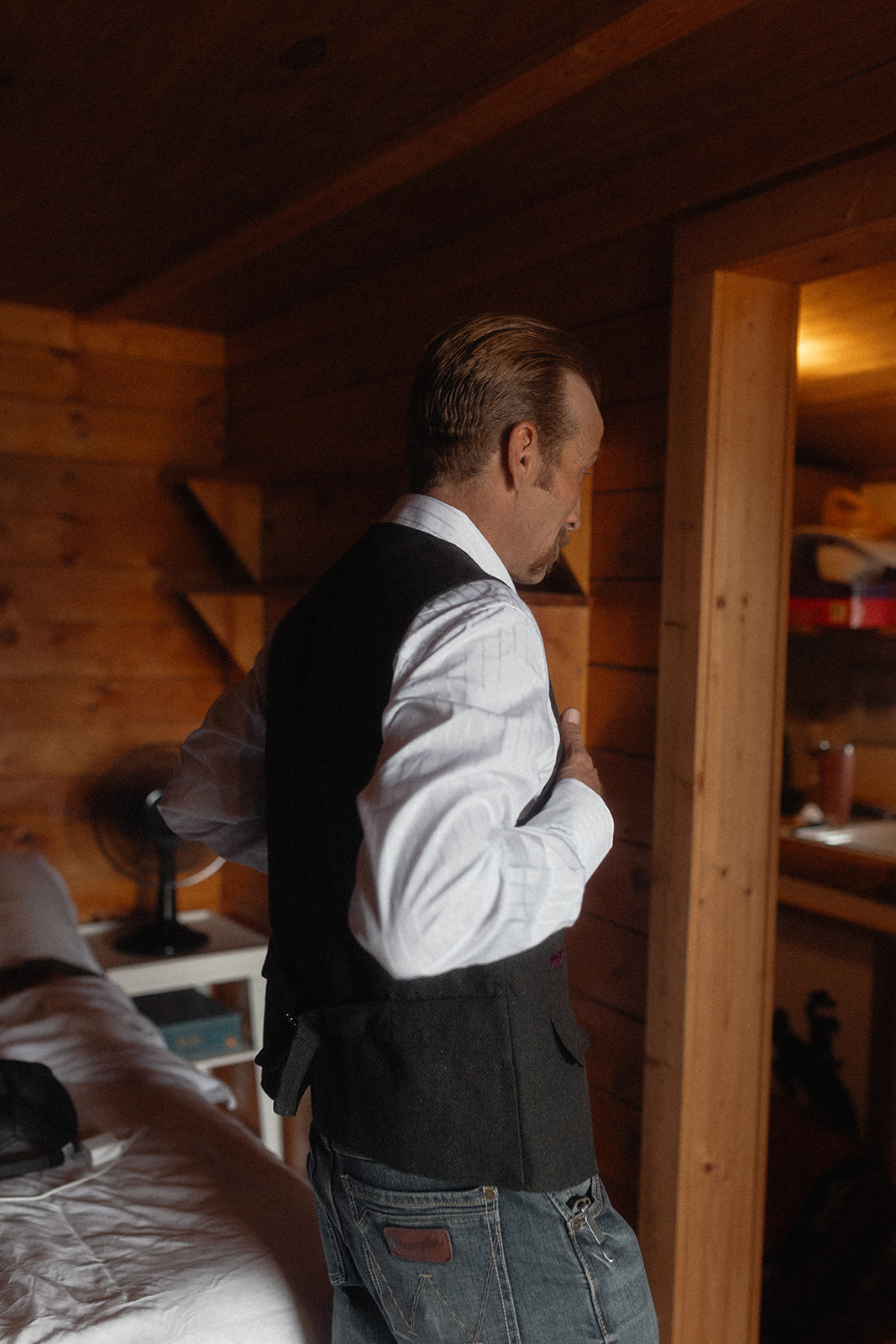 A person in a white shirt and dark vest adjusts their hair in front of a wooden wall mirror inside a rustic room for a wedding at preston ranch