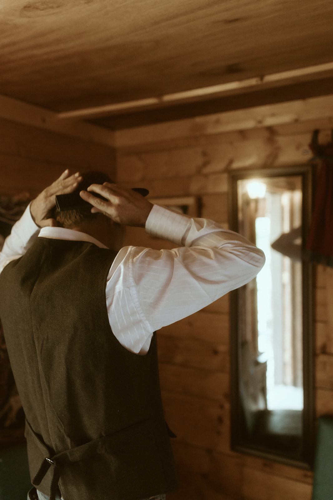 A person in a white shirt and dark vest adjusts their hair in front of a wooden wall mirror inside a rustic room for a wedding at preston ranch