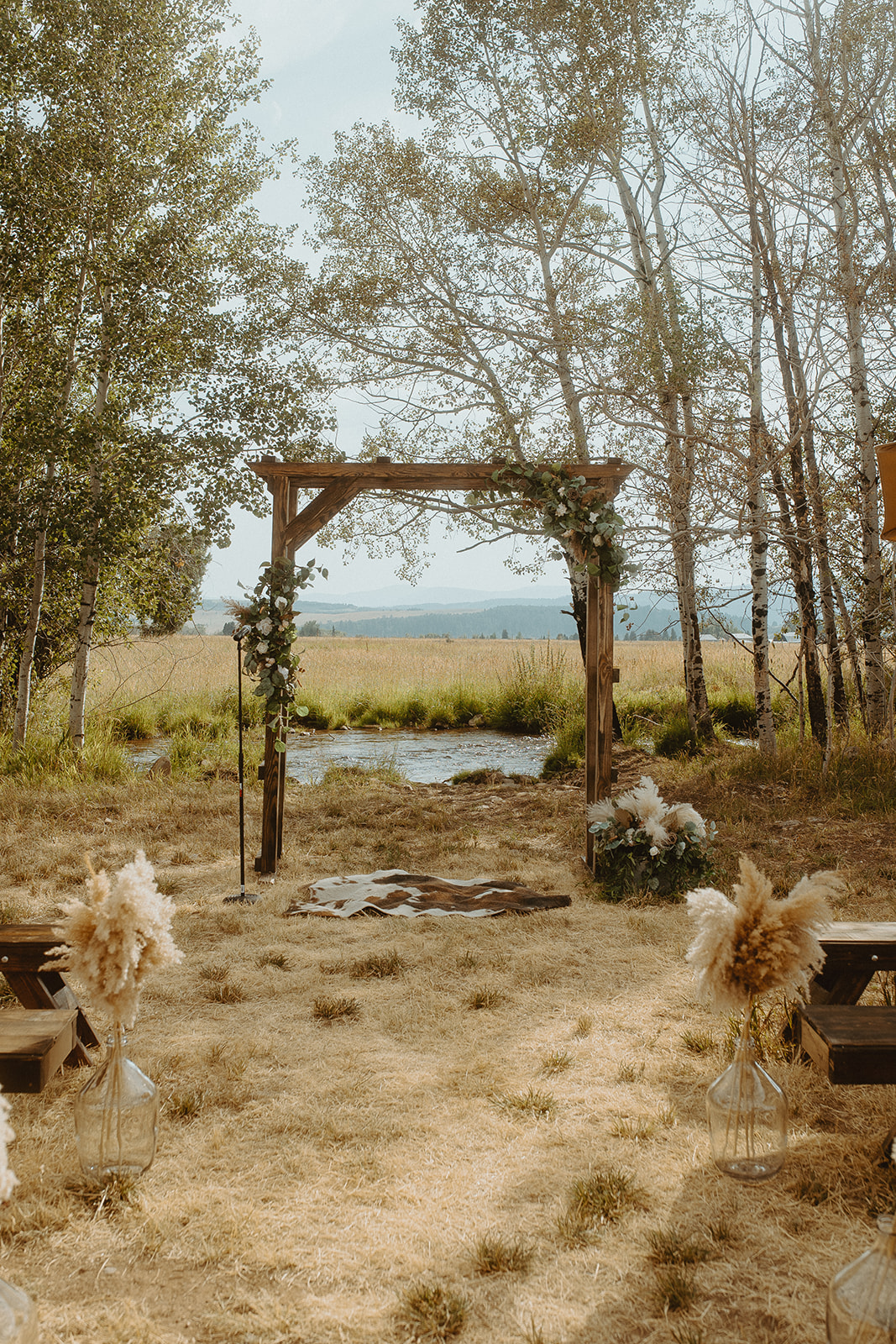 Outdoor wedding ceremony setup with a wooden arch adorned with flowers, surrounded by trees and grasses, overlooking a scenic landscape.