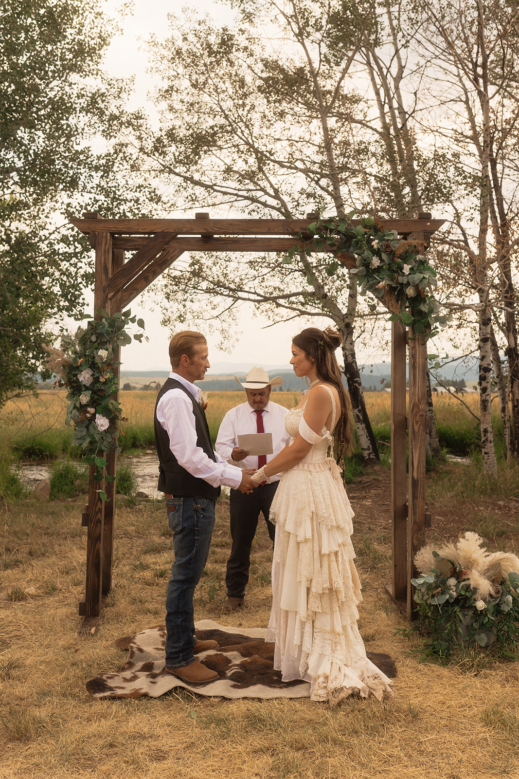 A couple stands under a wooden arch adorned with greenery and flowers, holding hands, while an officiant in a white hat and shirt reads from a book during an outdoor wedding ceremony.