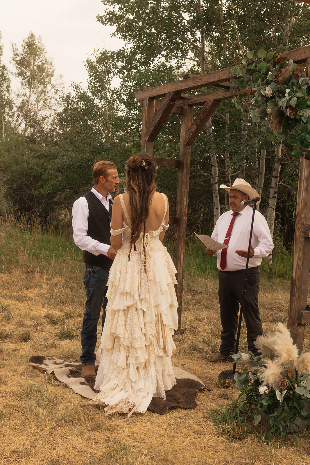 A couple stands under a wooden arch adorned with greenery and flowers, holding hands, while an officiant in a white hat and shirt reads from a book during an outdoor wedding ceremony.