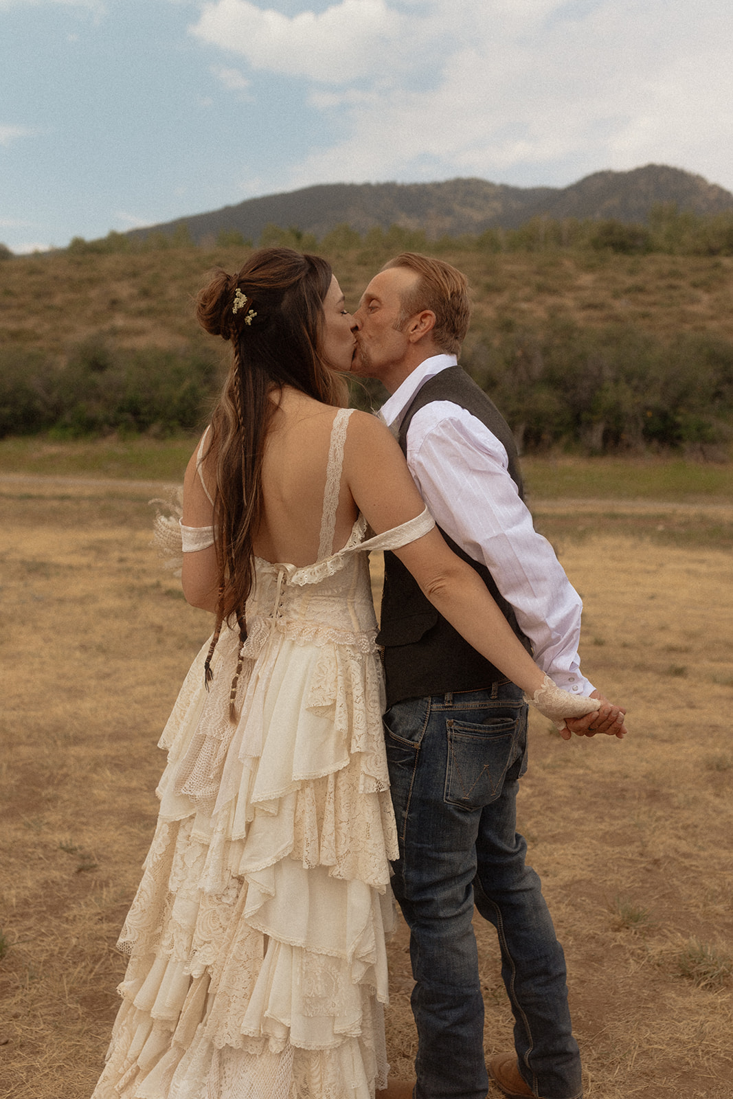 A couple dressed in wedding attire stands on a wooden bridge. The man, in a vest, embraces and kisses the woman, dressed in a flowing white gown. They are surrounded by a natural outdoor setting for their wedding at preston branch