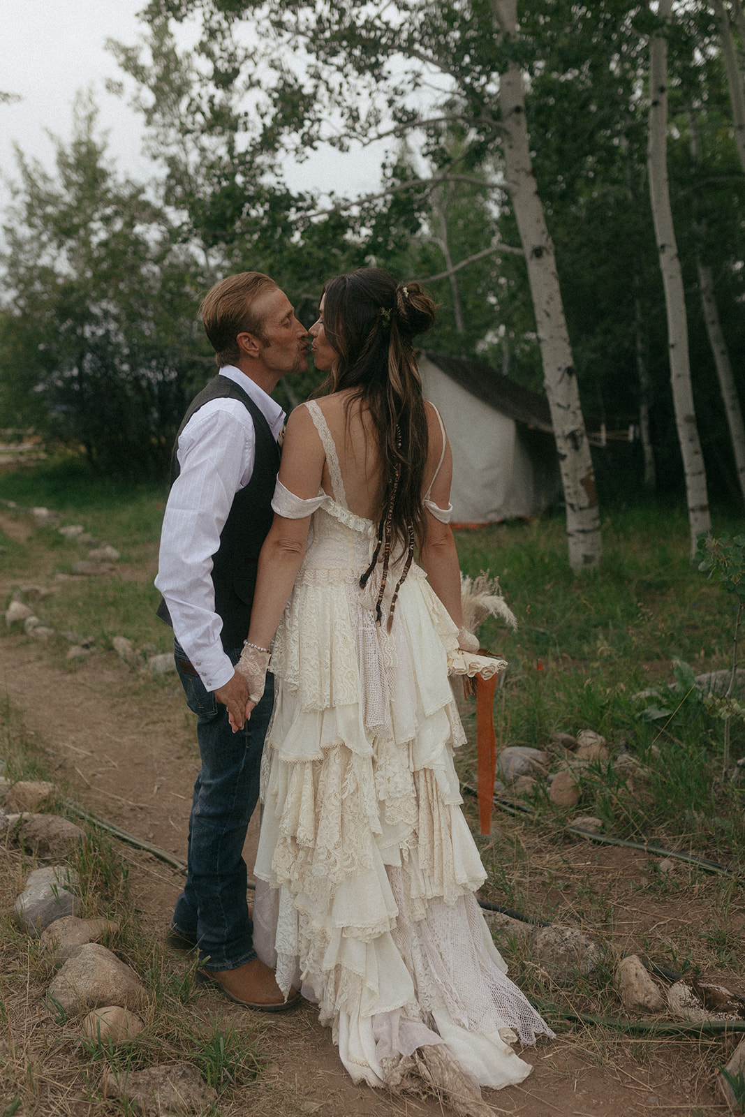 A couple dressed in wedding attire stands on a wooden bridge. The man, in a vest, embraces and kisses the woman, dressed in a flowing white gown. They are surrounded by a natural outdoor setting for their wedding at preston branch