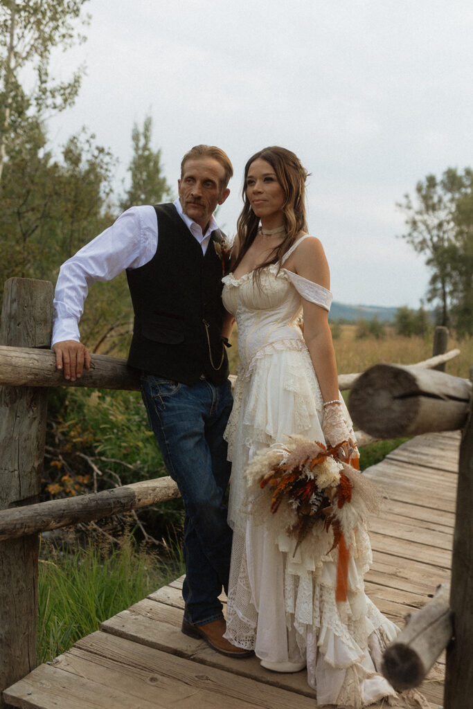 A couple dressed in wedding attire stands on a wooden bridge. The man, in a vest, embraces and kisses the woman, dressed in a flowing white gown. They are surrounded by a natural outdoor setting for their wedding at preston branch