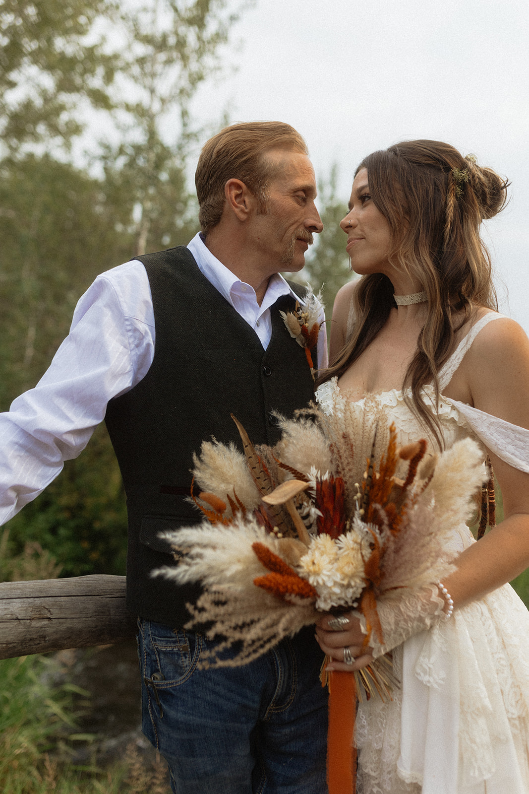 A couple dressed in wedding attire stands on a wooden bridge. The man, in a vest, embraces and kisses the woman, dressed in a flowing white gown. They are surrounded by a natural outdoor setting for their wedding at preston branch