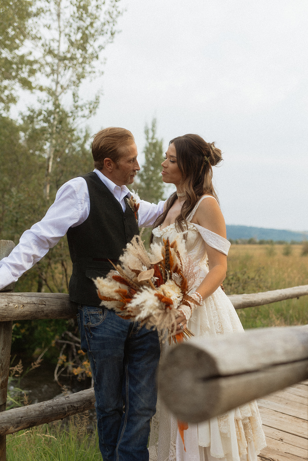 A couple dressed in wedding attire stands on a wooden bridge. The man, in a vest, embraces and kisses the woman, dressed in a flowing white gown. They are surrounded by a natural outdoor setting for their wedding at preston branch