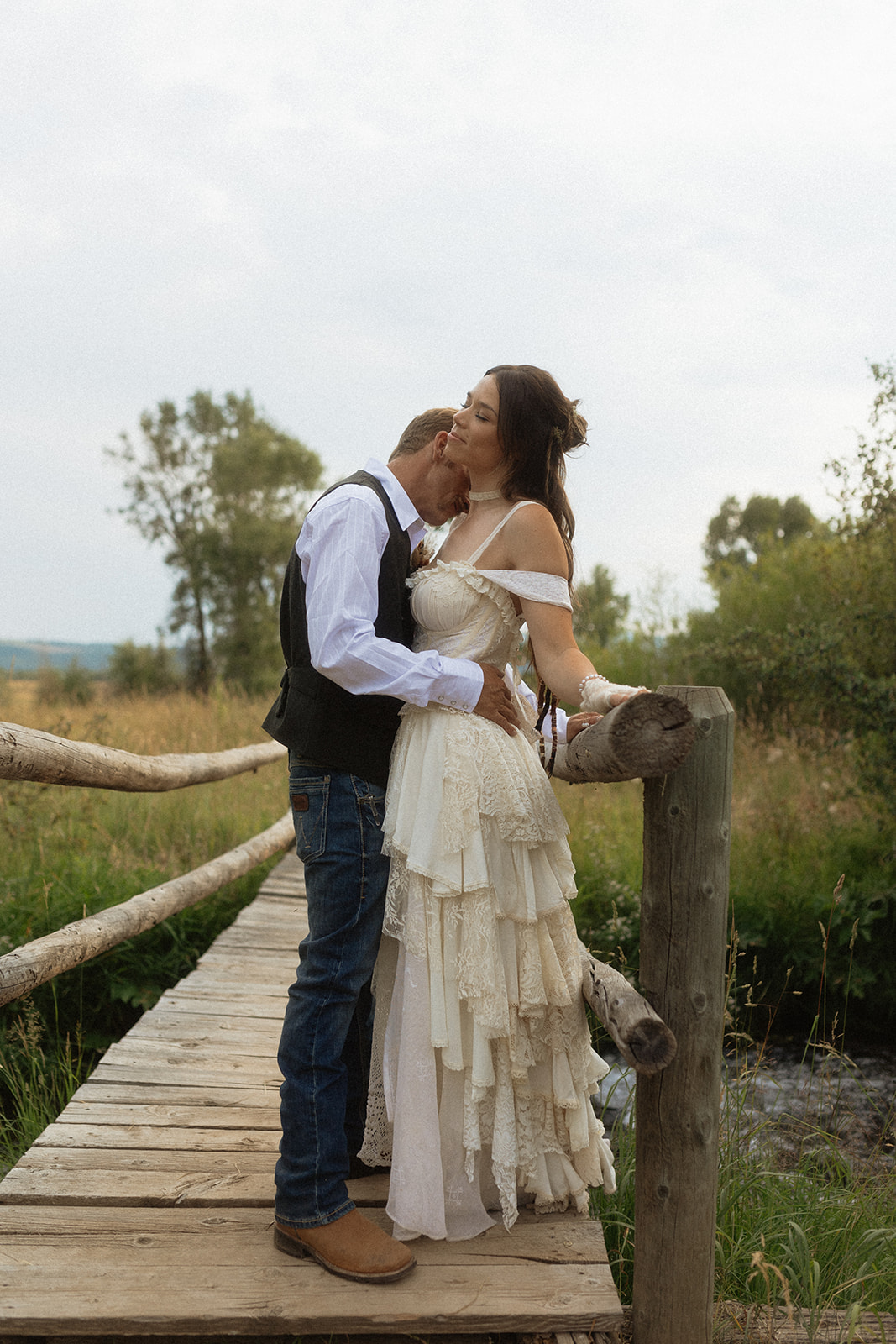 A couple dressed in wedding attire stands on a wooden bridge. The man, in a vest, embraces and kisses the woman, dressed in a flowing white gown. They are surrounded by a natural outdoor setting for their wedding at preston branch