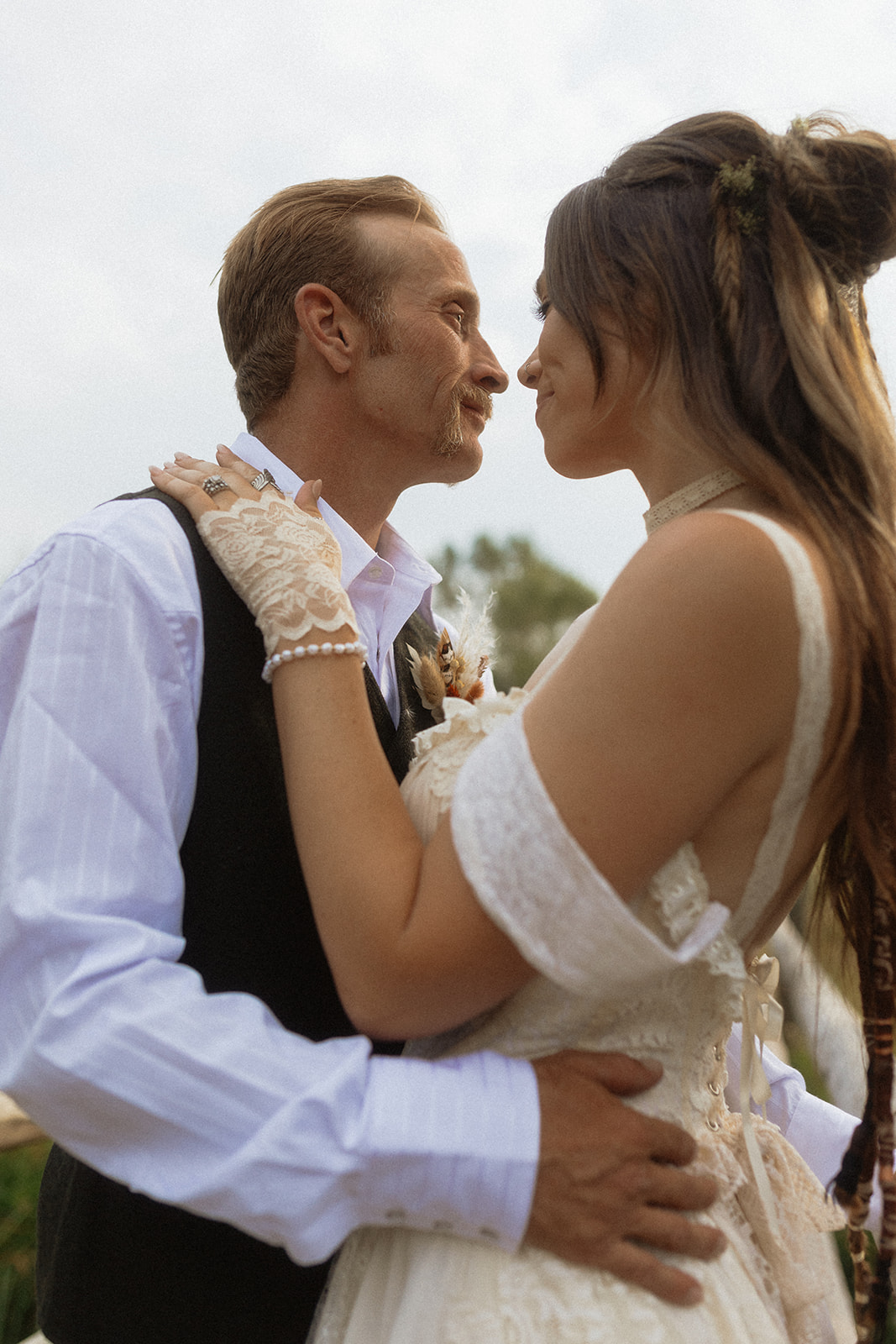 A couple dressed in wedding attire stands on a wooden bridge. The man, in a vest, embraces and kisses the woman, dressed in a flowing white gown. They are surrounded by a natural outdoor setting for their wedding at preston branch