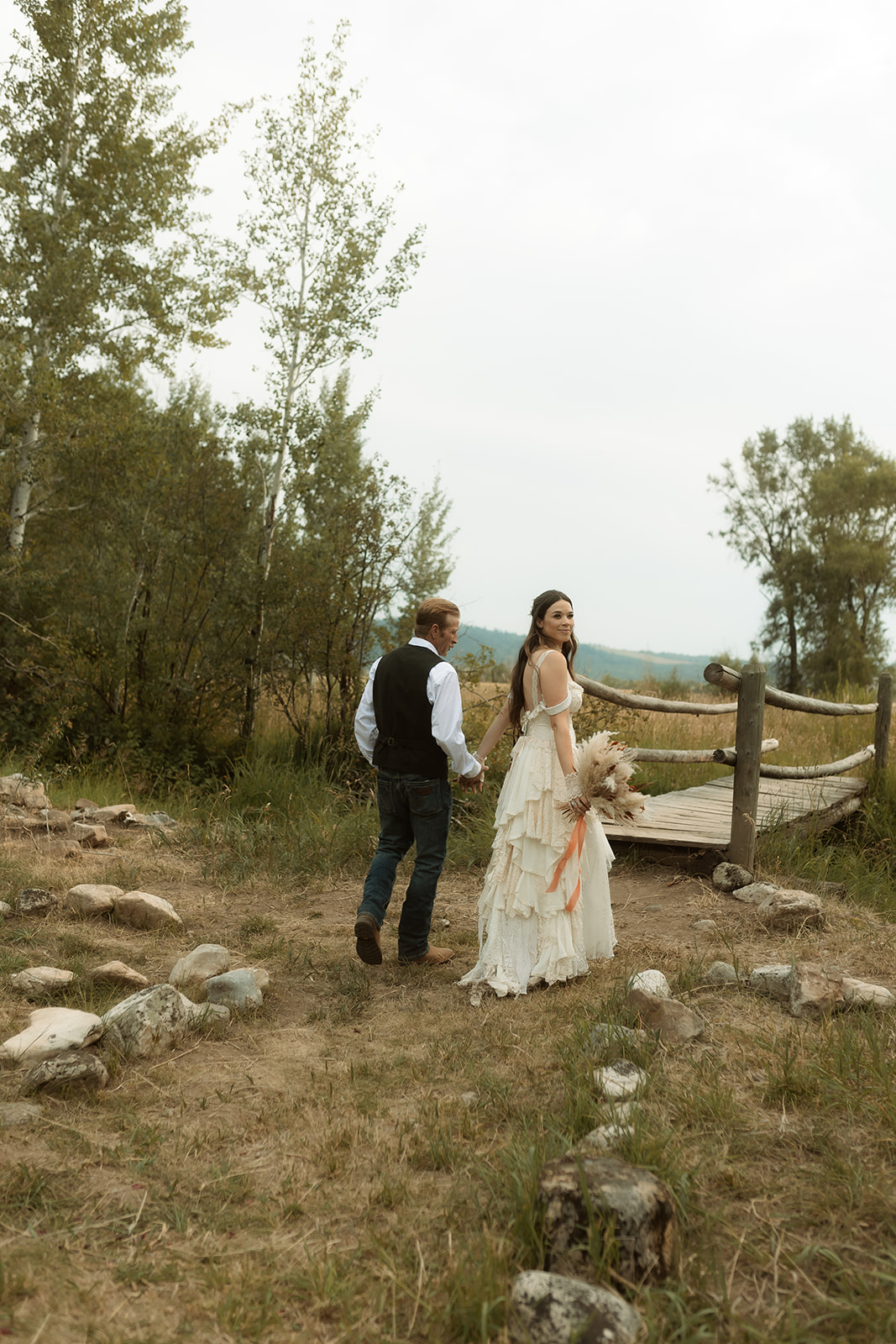 A couple dressed in wedding attire stands on a wooden bridge. The man, in a vest, embraces and kisses the woman, dressed in a flowing white gown. They are surrounded by a natural outdoor setting for their wedding at preston branch