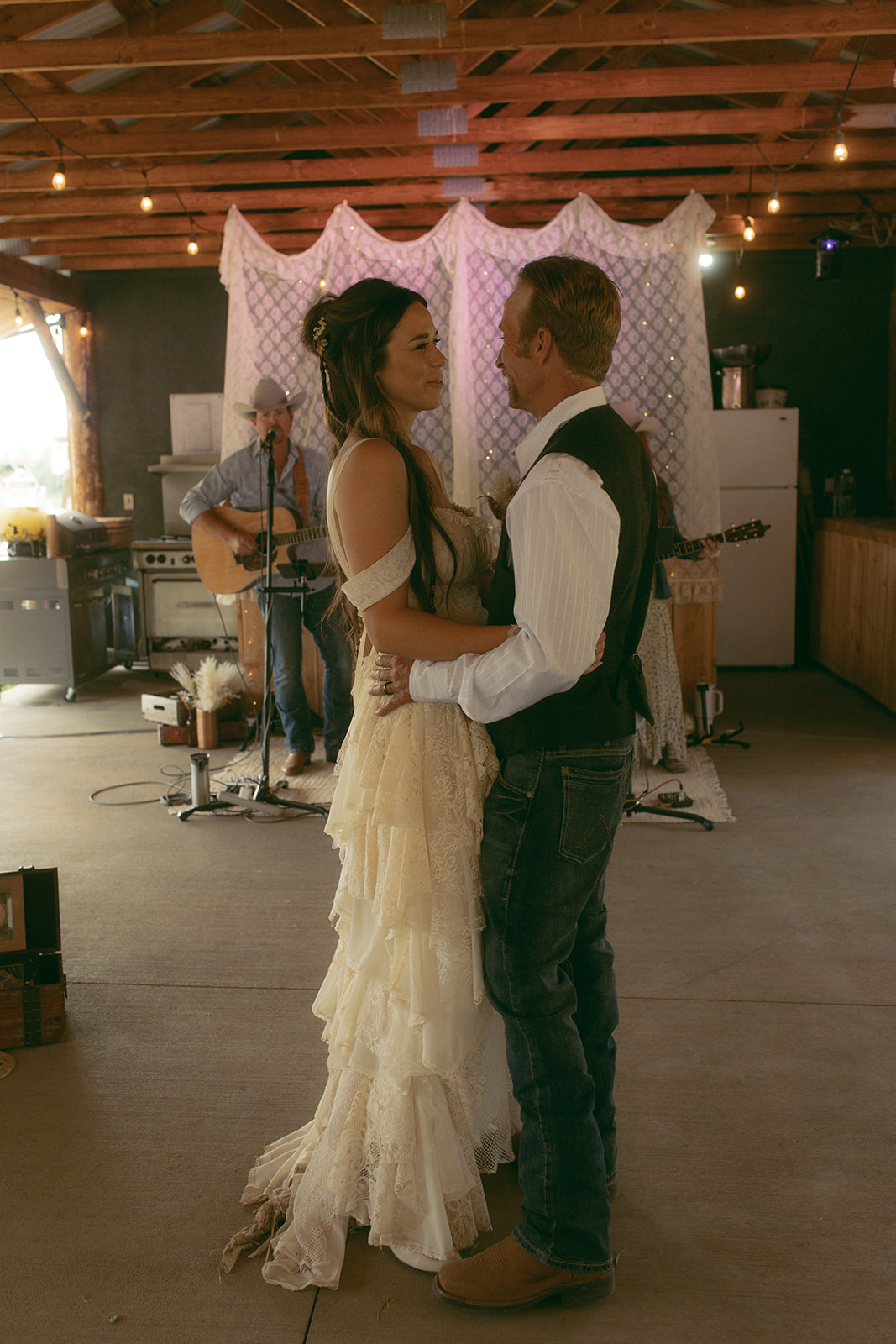 A couple dances in a rustic setting with string lights overhead. A musician plays guitar in the background. The woman wears a white dress and the man a vest as they have their first dance at preston ranch