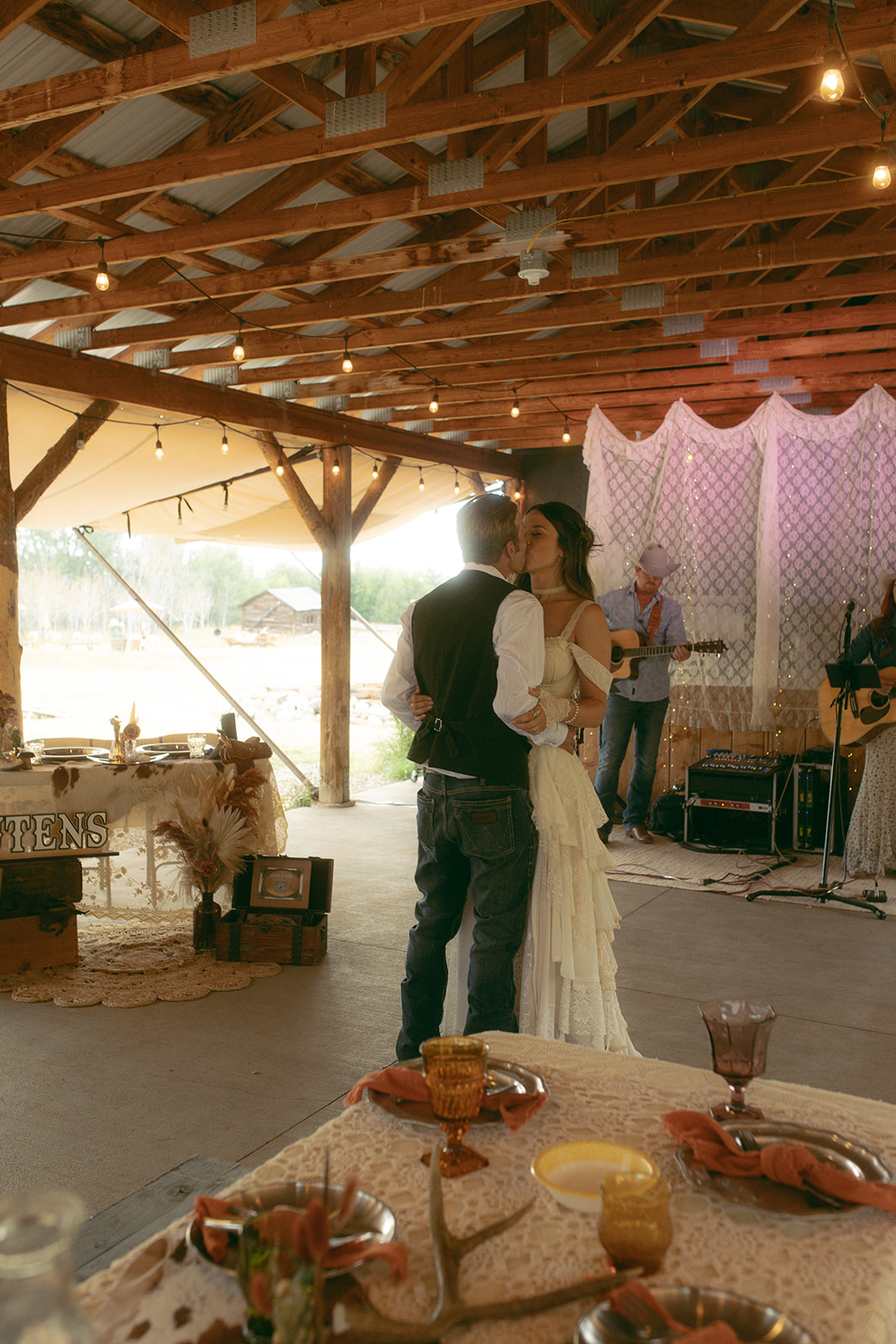 A couple dances in a rustic setting with string lights overhead. A musician plays guitar in the background. The woman wears a white dress and the man a vest as they have their first dance at preston ranch