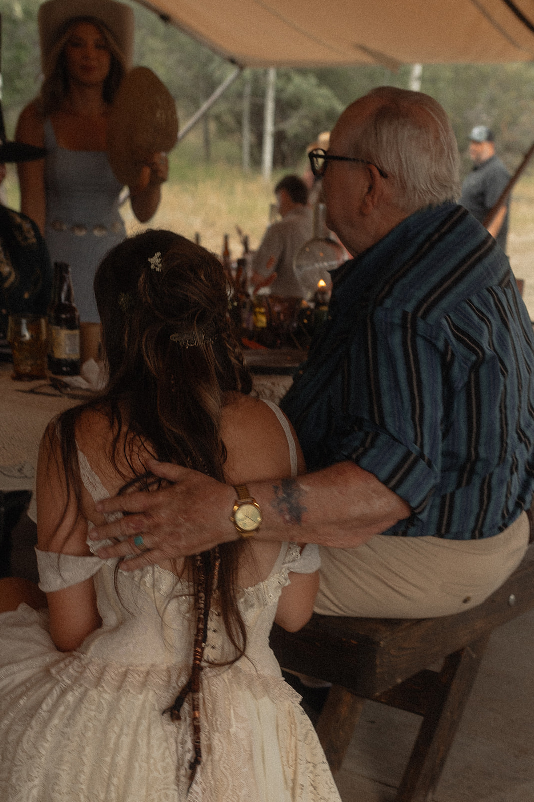 An elderly man with glasses has his arm around a young woman in a white dress. They are seated at an outdoor event under a tent. Other people and tables with items can be seen in the background.