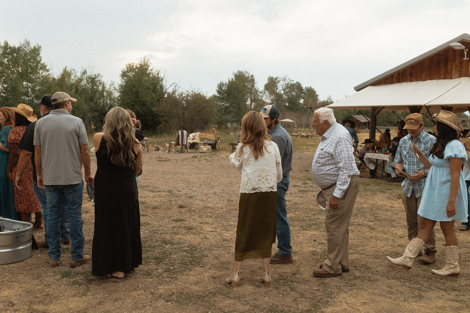 A group of people stands outside in a rural setting near a building with a wooden roof. They are casually dressed, some in hats, engaging in conversation. Trees and distant people are visible in the background.