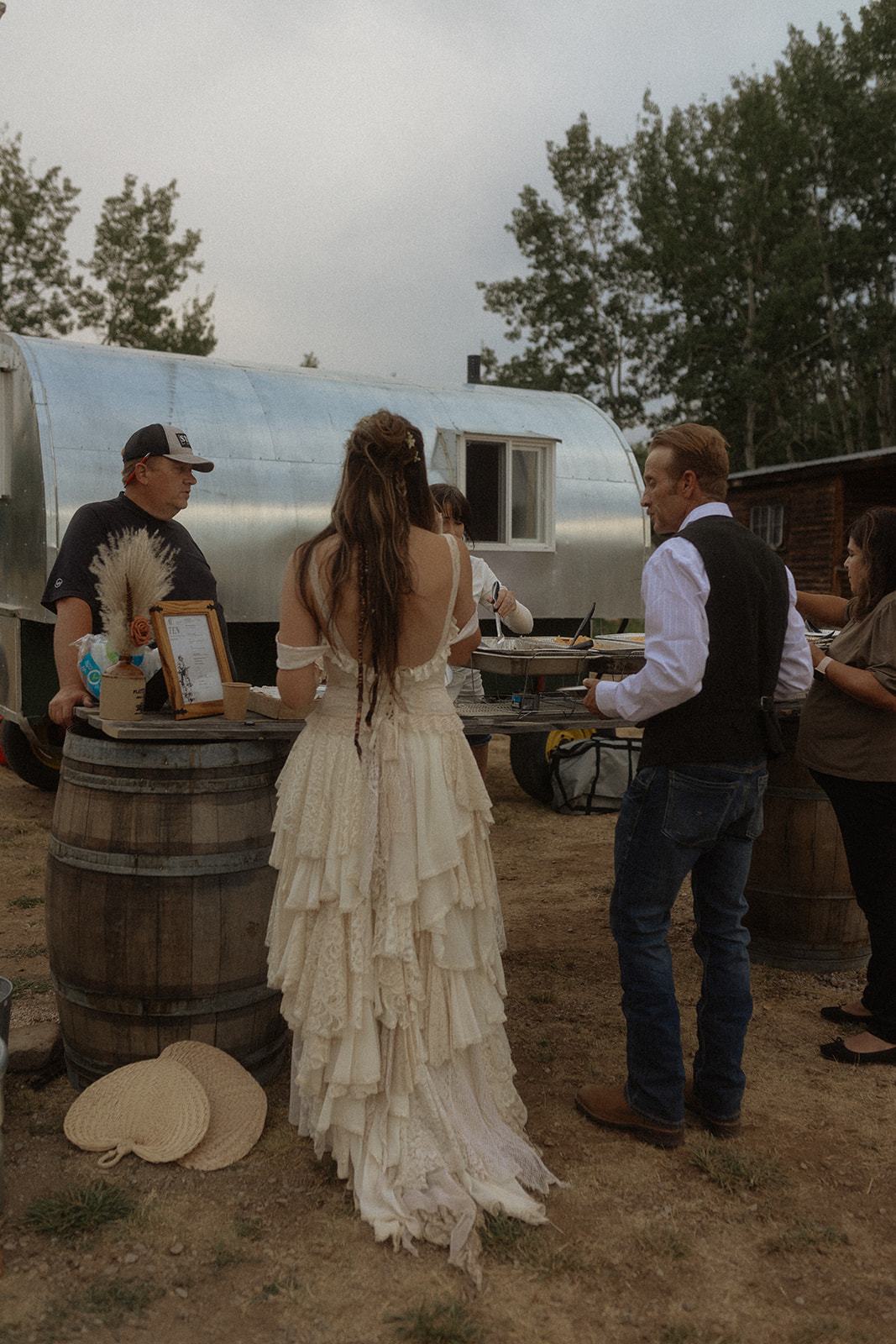 A bride and groom stand at an outdoor serving station near a silver trailer, with the bride in a tiered dress and the groom in a vest and jeans. A server in casual attire and another person are also present for their wedding at preston ranch