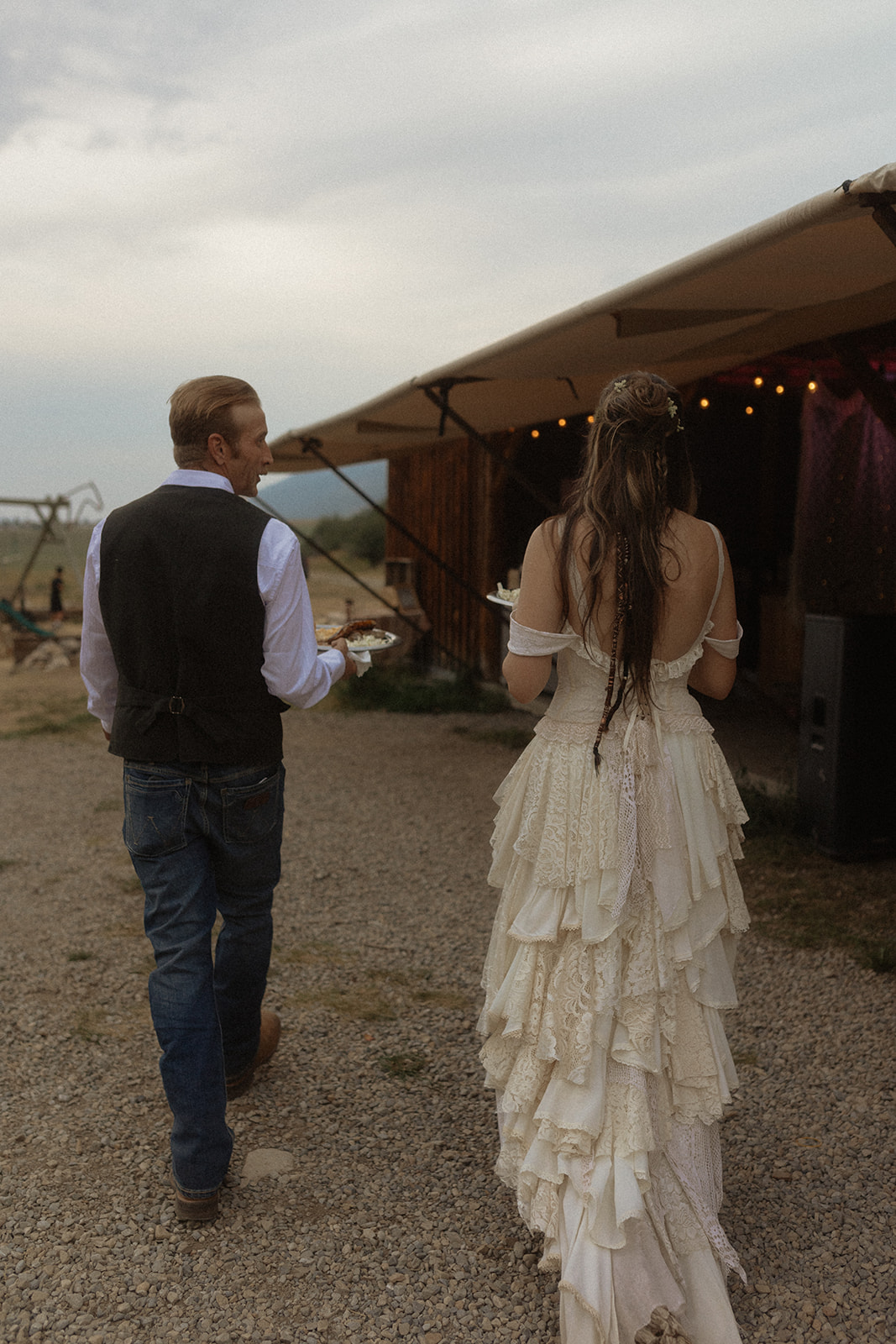 A bride and groom stand at an outdoor serving station near a silver trailer, with the bride in a tiered dress and the groom in a vest and jeans. A server in casual attire and another person are also present for their wedding at preston ranch 