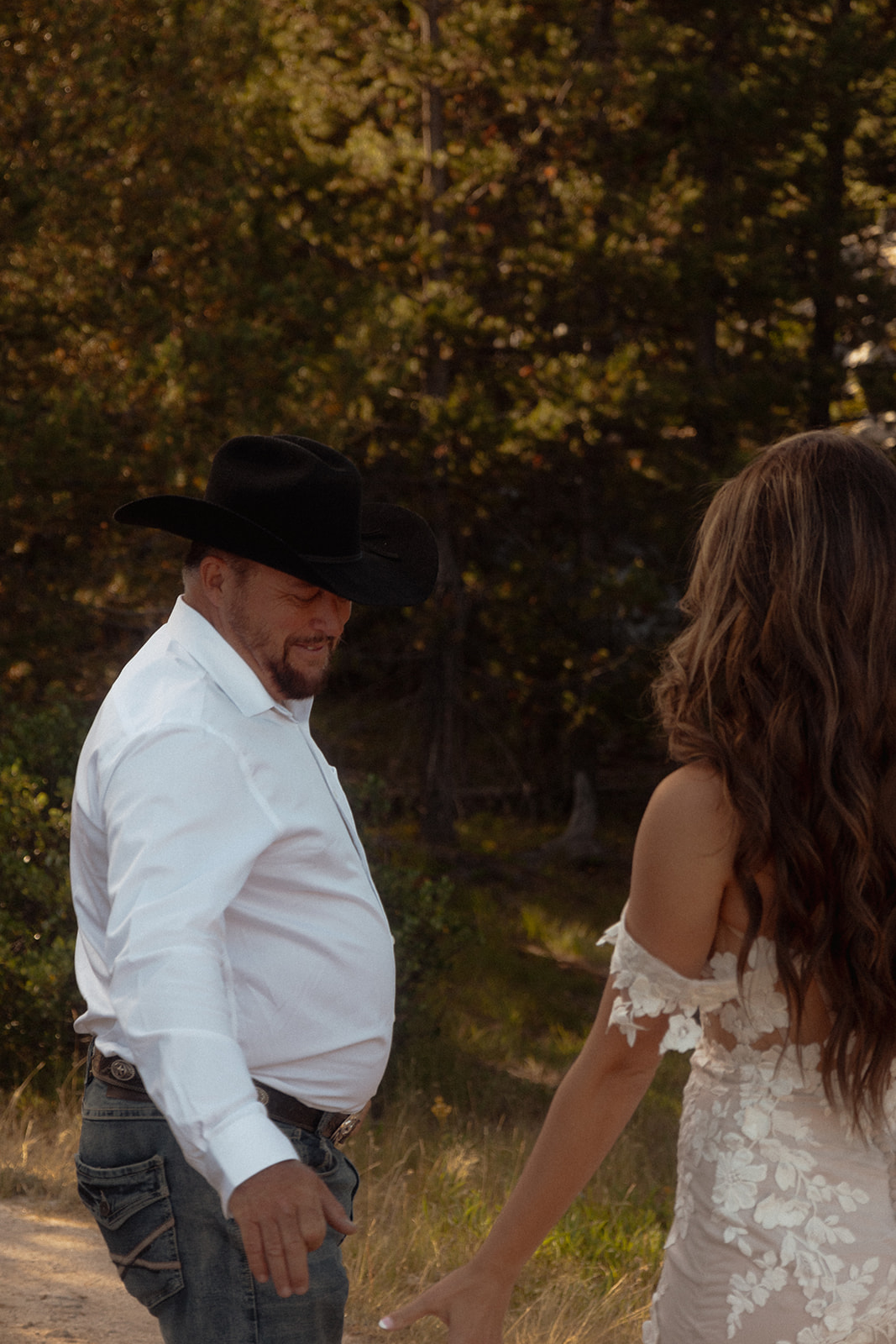 A man in a white shirt and black cowboy hat stands outdoors with arms outstretched, facing a woman in a white dress. Trees are visible in the background for an Grand Teton National park elopement at taggart lake