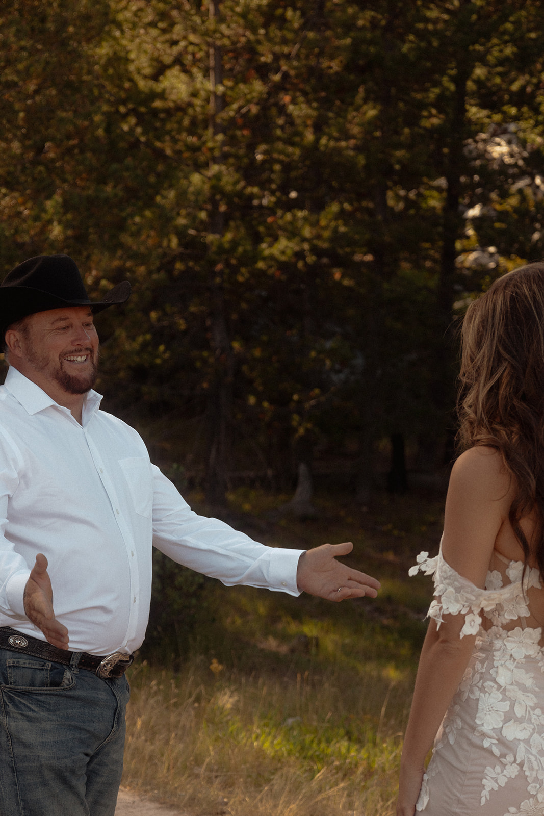 A man in a white shirt and black cowboy hat stands outdoors with arms outstretched, facing a woman in a white dress. Trees are visible in the background for an  Grand Teton National park elopement at taggart lake