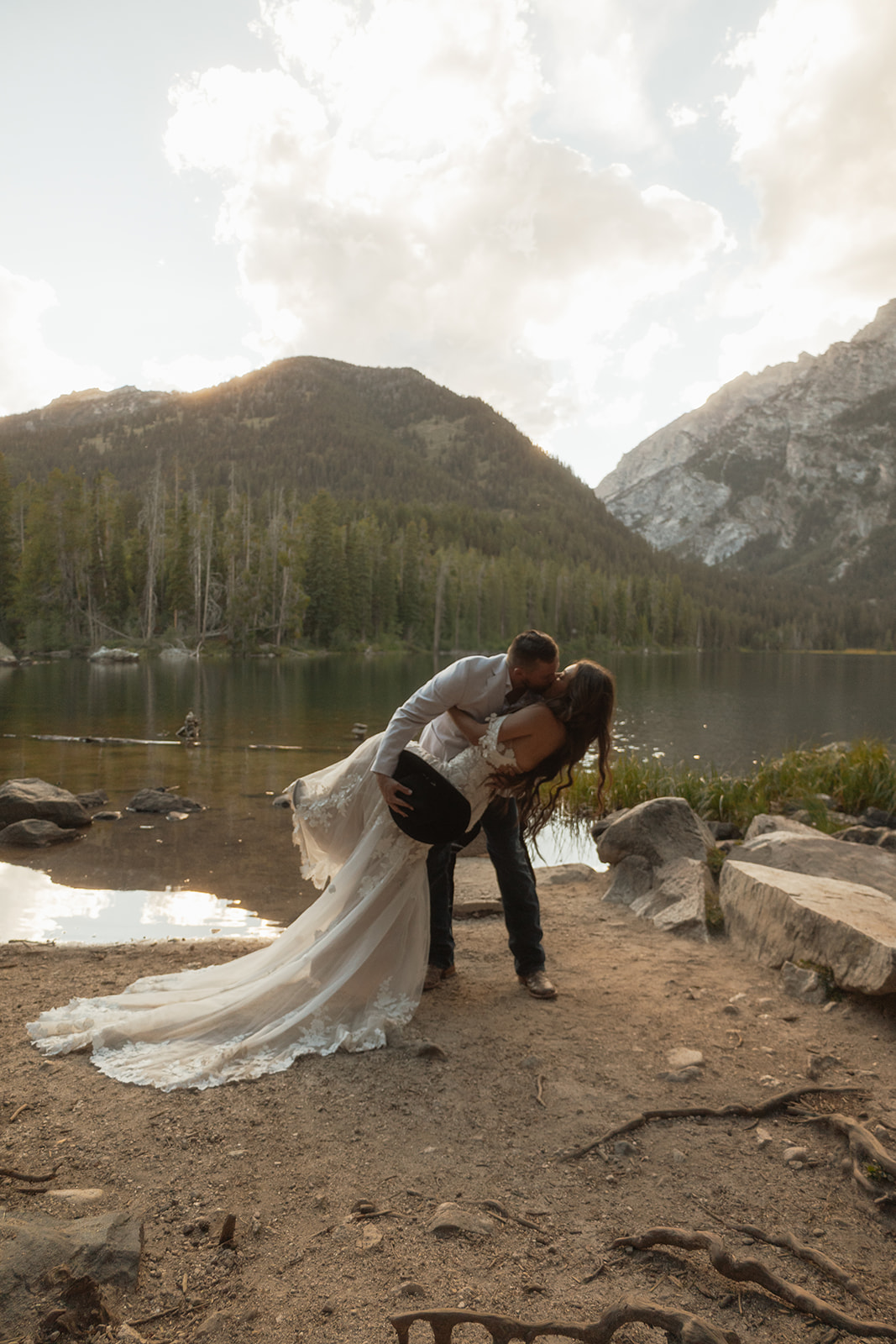 A couple in wedding attire shares a kiss by a lake with mountains and trees in the background for a Grand Teton National park at taggart lake