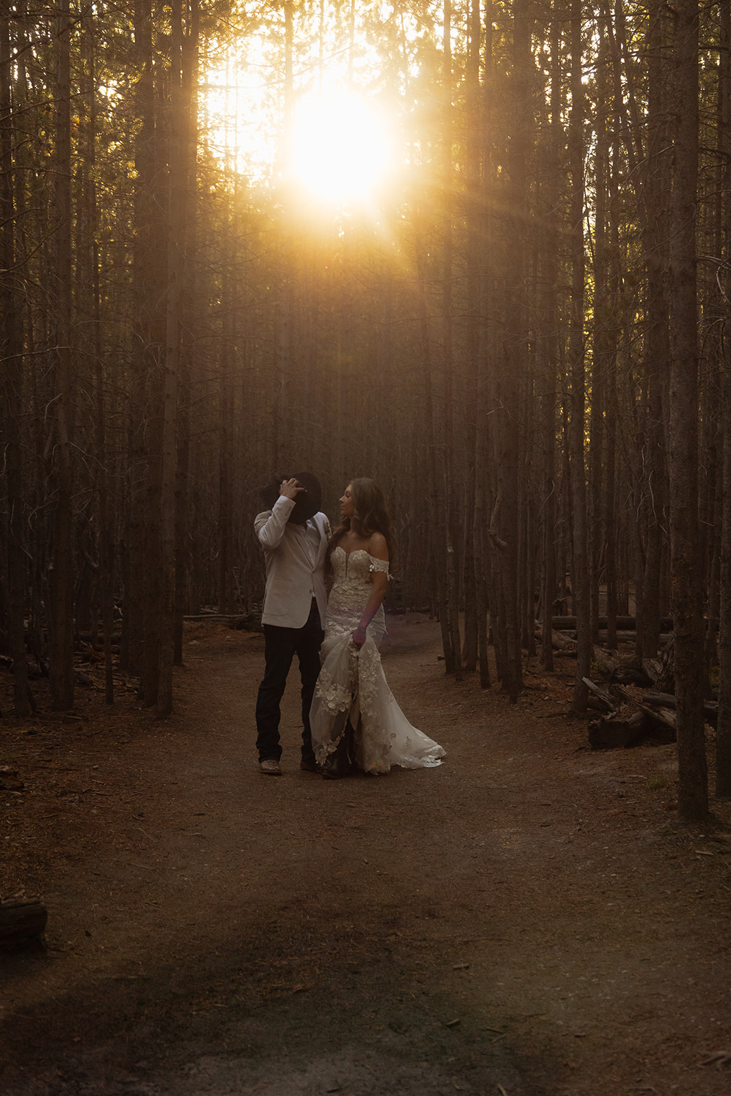 A couple dressed in wedding attire stands in the middle of a forest path with sunlight filtering through the trees 