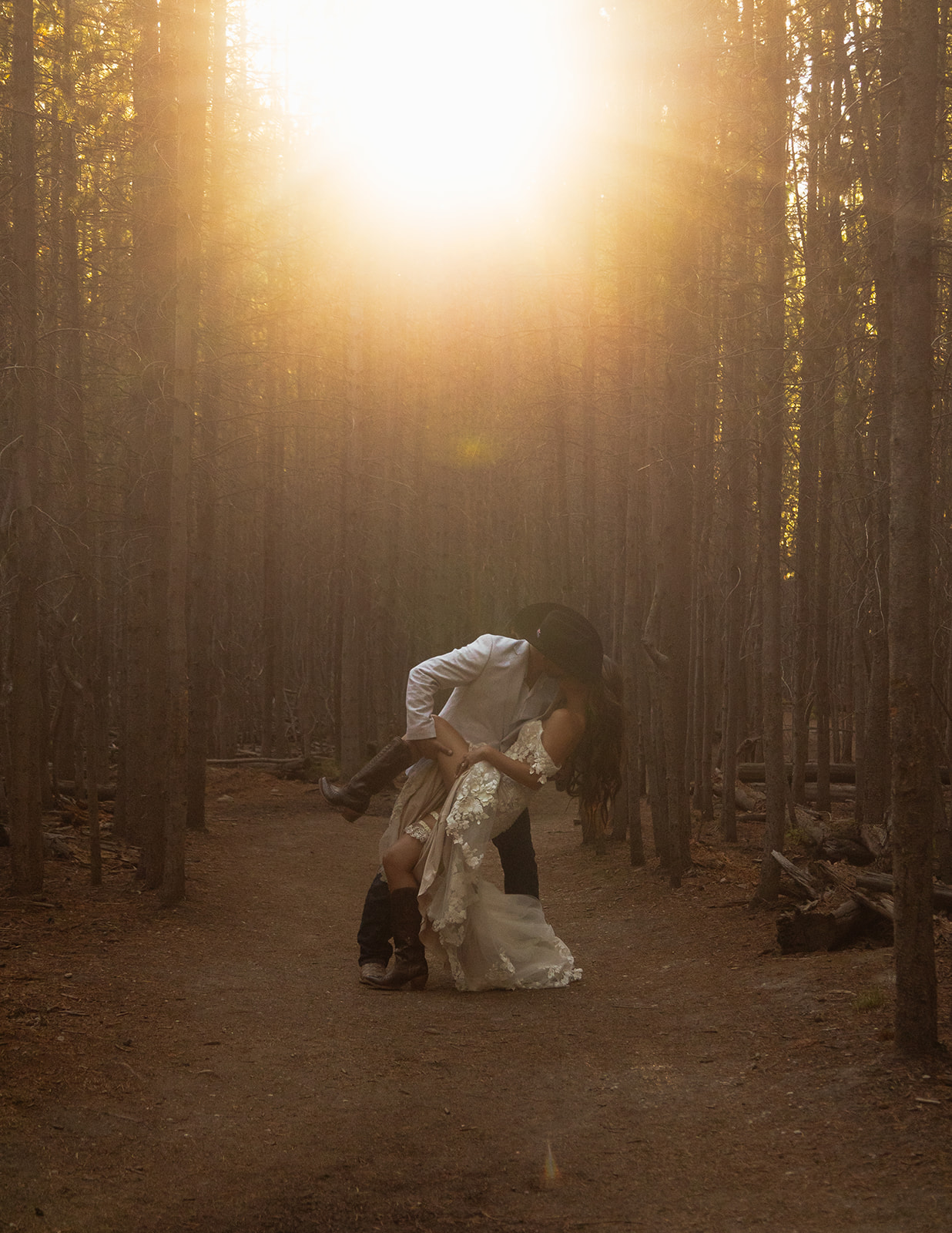 A couple dressed in wedding attire stands in the middle of a forest path with sunlight filtering through the trees 