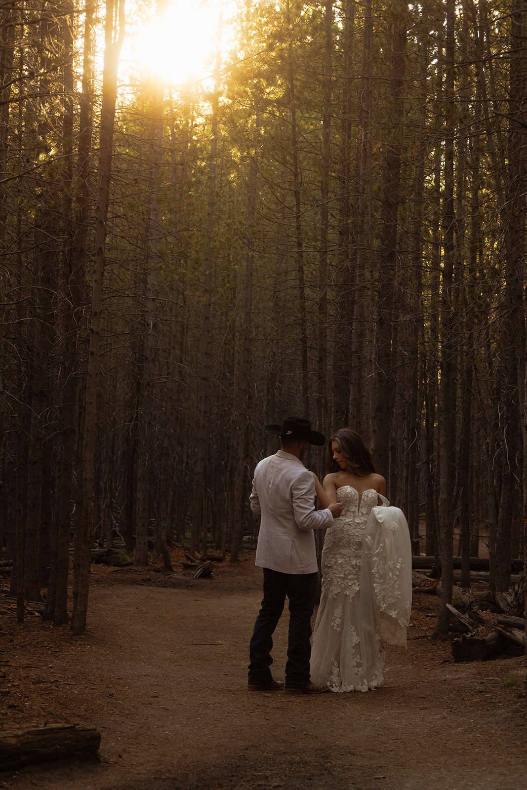 A couple dressed in wedding attire stands in the middle of a forest path with sunlight filtering through the trees 