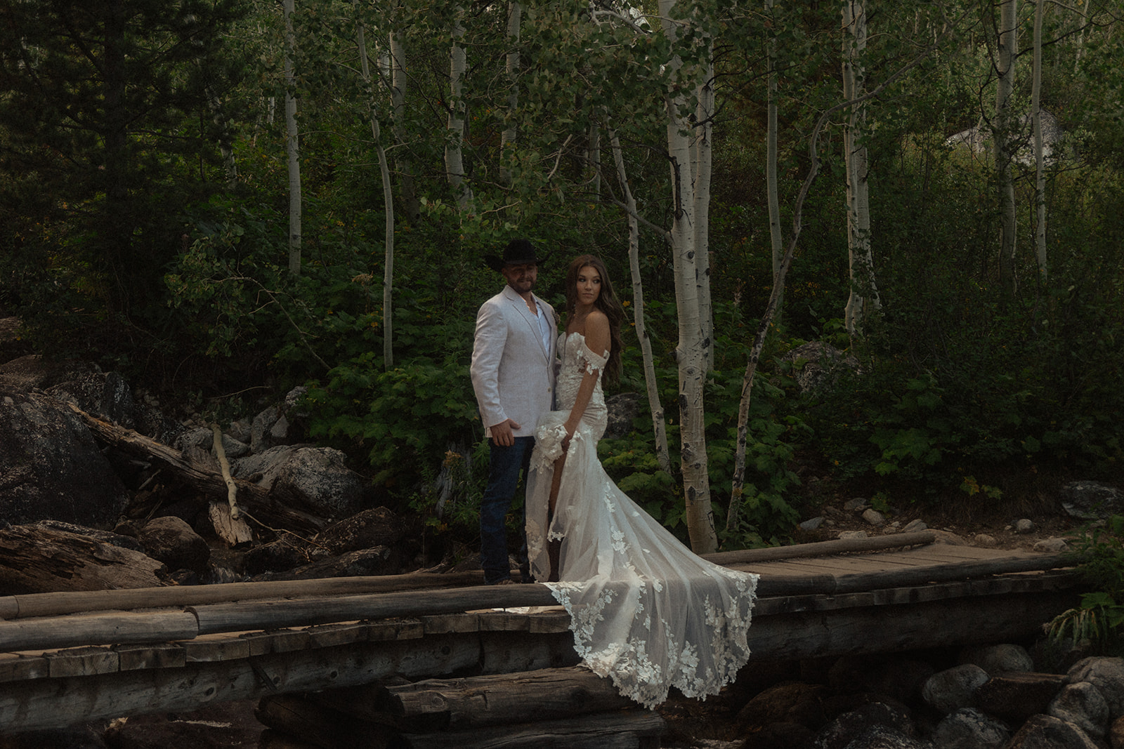 A couple stands on a wooden bridge in a forested area. The woman wears a long, white lace gown while the man wears a white jacket and dark pants. Trees and a stream are visible in the background for a Grand Teton National park taggart lake elopement