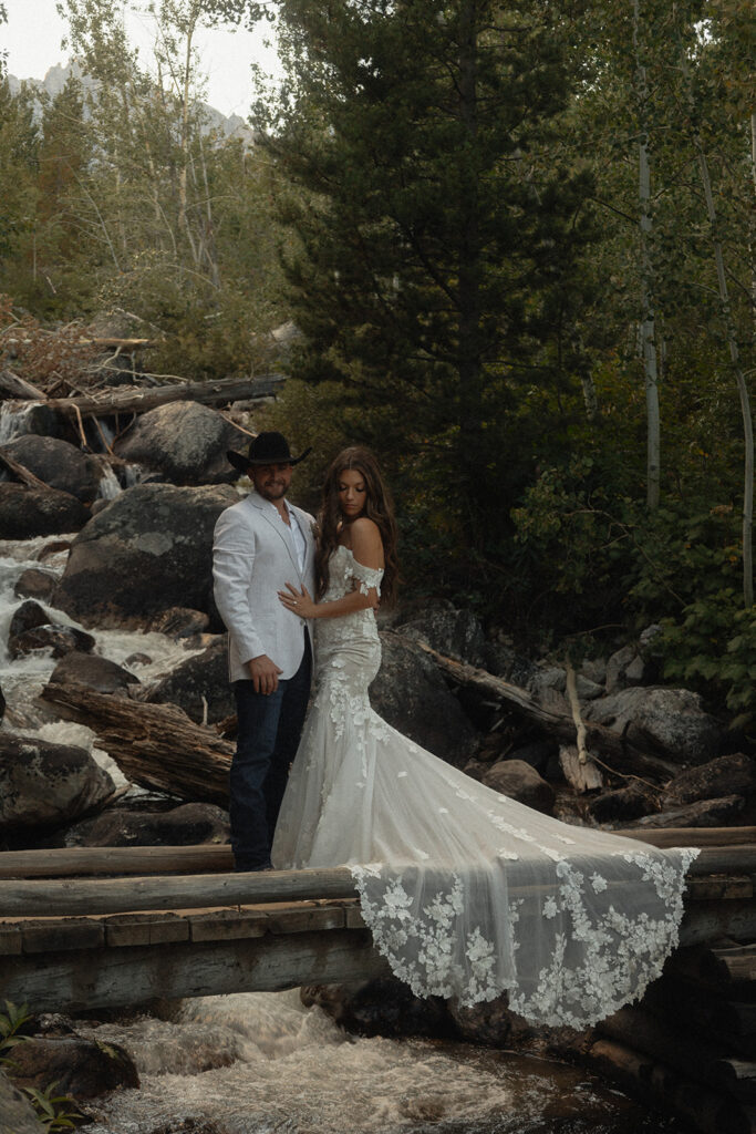 A couple stands on a wooden bridge in a forested area. The woman wears a long, white lace gown while the man wears a white jacket and dark pants. Trees and a stream are visible in the background for a Grand Teton National park taggart lake elopement